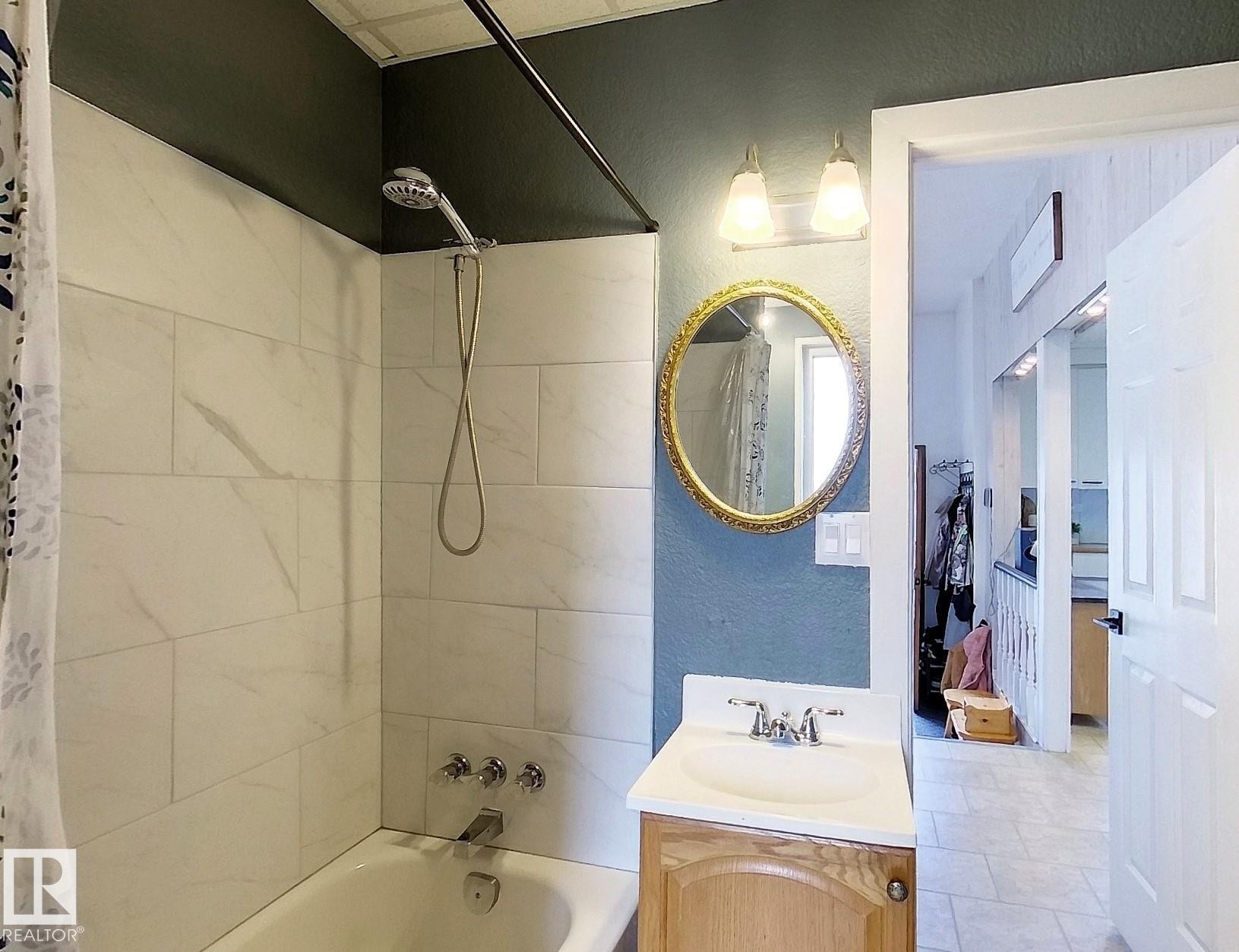 Bathroom featuring a classic tub and shower combination with white subway tile surround, a single vanity with a light wood-finish cabinet, and a decorative round mirror - 7126 4A Ave, Edson, AB - Indoor Photo Showing Bathroom