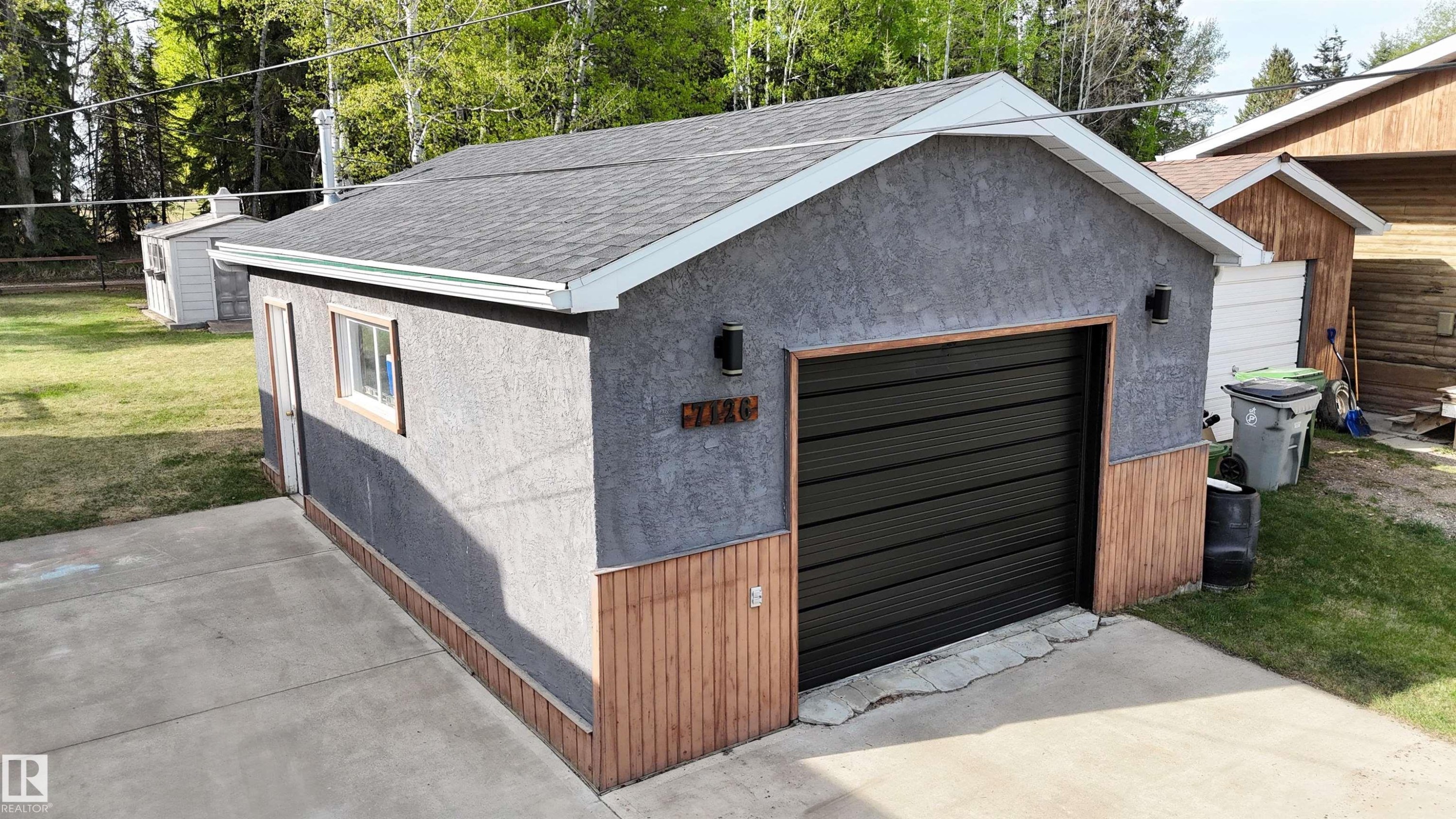 Detached garage featuring a dark gray roll-up door, stucco siding, wood-finish paneling, and two exterior light fixtures - 7126 4A Ave, Edson, AB - Outdoor With Exterior