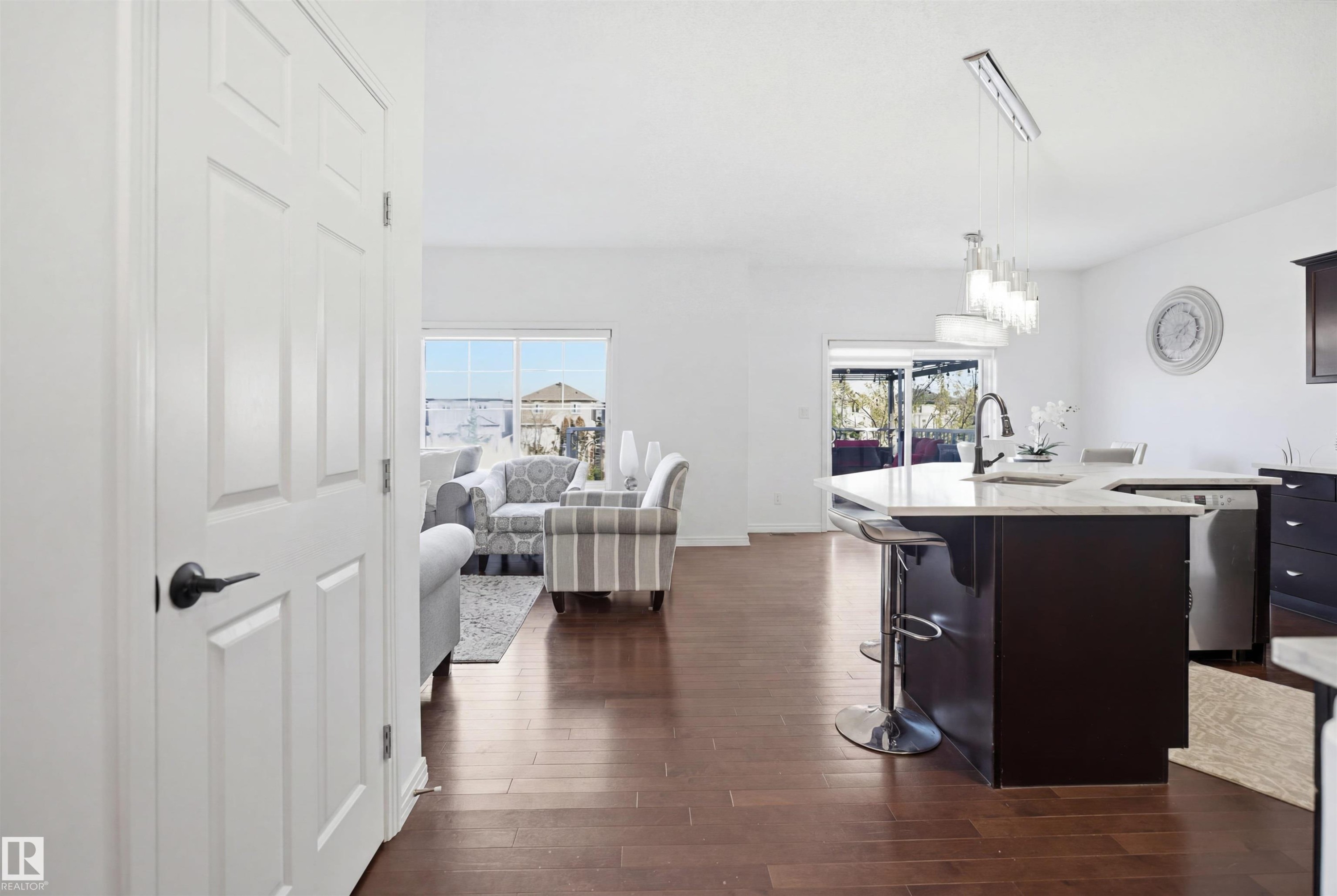 Open-concept interior featuring wood-finish flooring, a kitchen island with stone countertops, a modern linear pendant light fixture, and white paneled interior doors with dark hardware - 1453 Hays Way Nw, Edmonton, AB - Indoor Photo Showing Kitchen