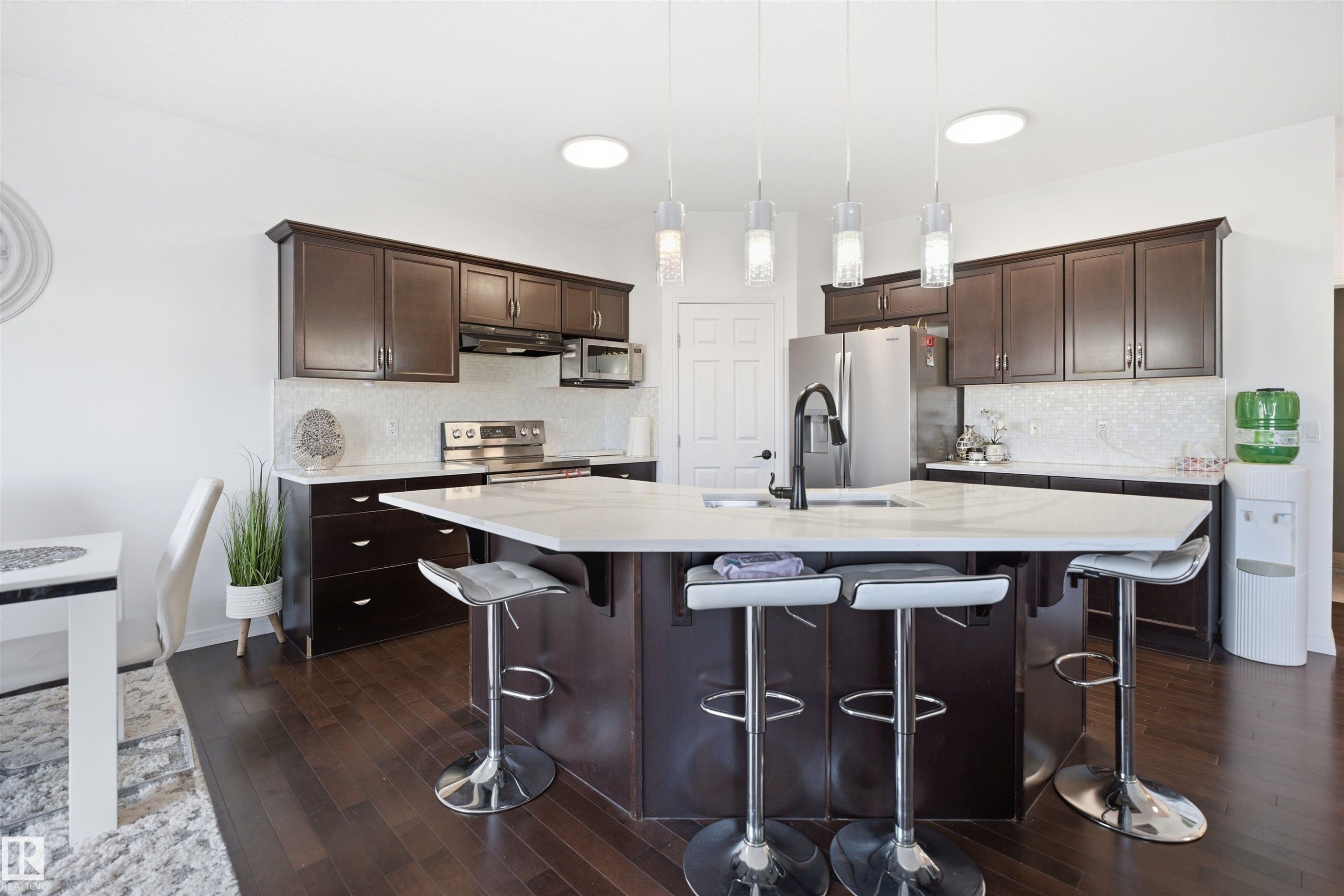 Kitchen featuring dark wood cabinetry, a white countertop island with seating, a mosaic tile backsplash, stainless steel appliances, and dark wood-finish flooring - 1453 Hays Way Nw, Edmonton, AB - Indoor Photo Showing Kitchen With Stainless Steel Kitchen With Upgraded Kitchen