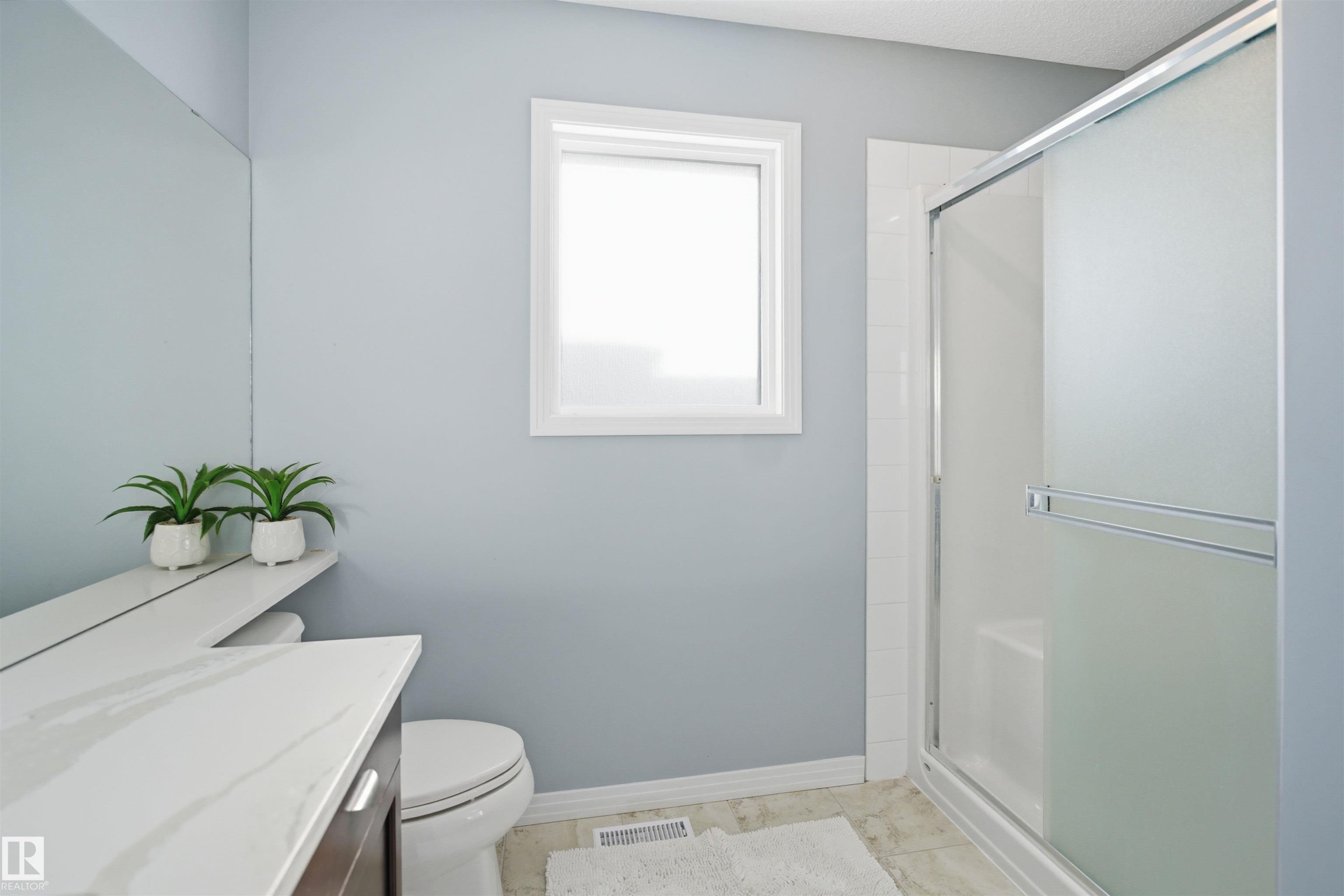 Bathroom featuring a frosted glass shower enclosure, white window frame, wood-finish vanity with a light-toned countertop, and light blue wall paint - 1453 Hays Way Nw, Edmonton, AB - Indoor Photo Showing Bathroom