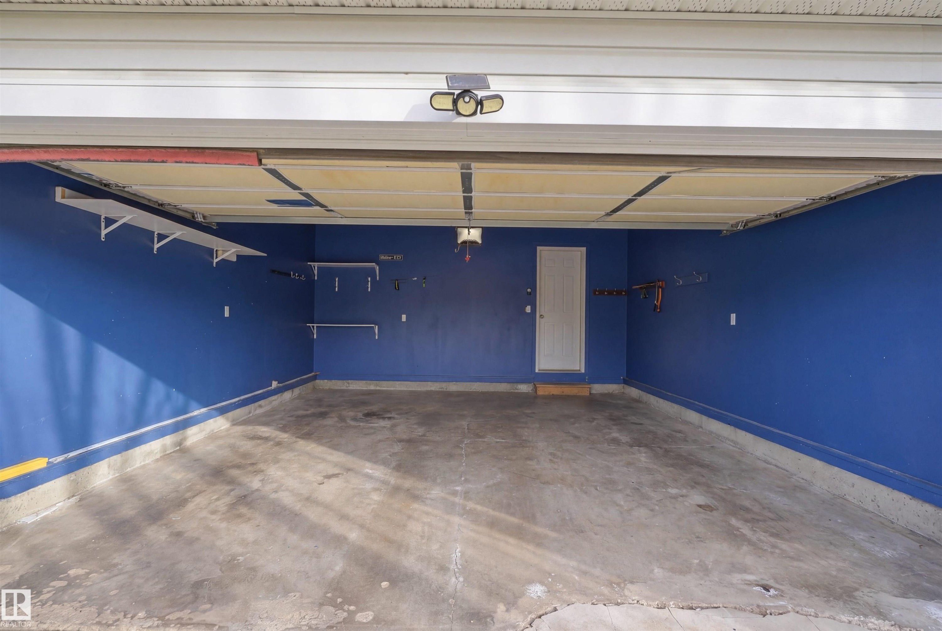 Spacious garage featuring a concrete floor, vibrant blue walls, dedicated side door access, and multiple built-in shelving units for organized storage - 1453 Hays Way Nw, Edmonton, AB - Indoor Photo Showing Garage