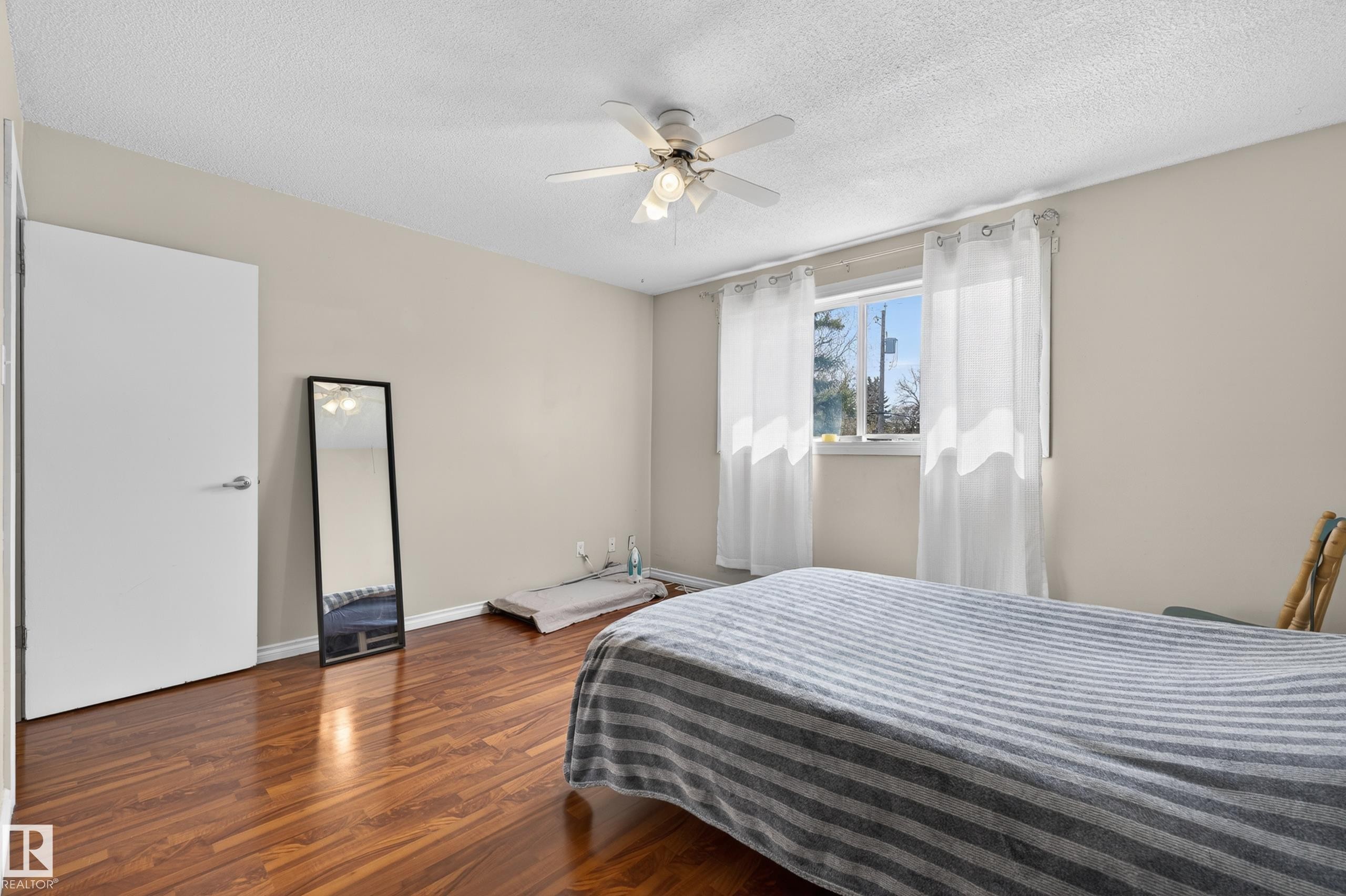 Room featuring wood-finish flooring, a ceiling fan with integrated lighting, and a window with white sheer curtains - 26E Meadowlark Village, Edmonton, AB - Indoor Photo Showing Bedroom