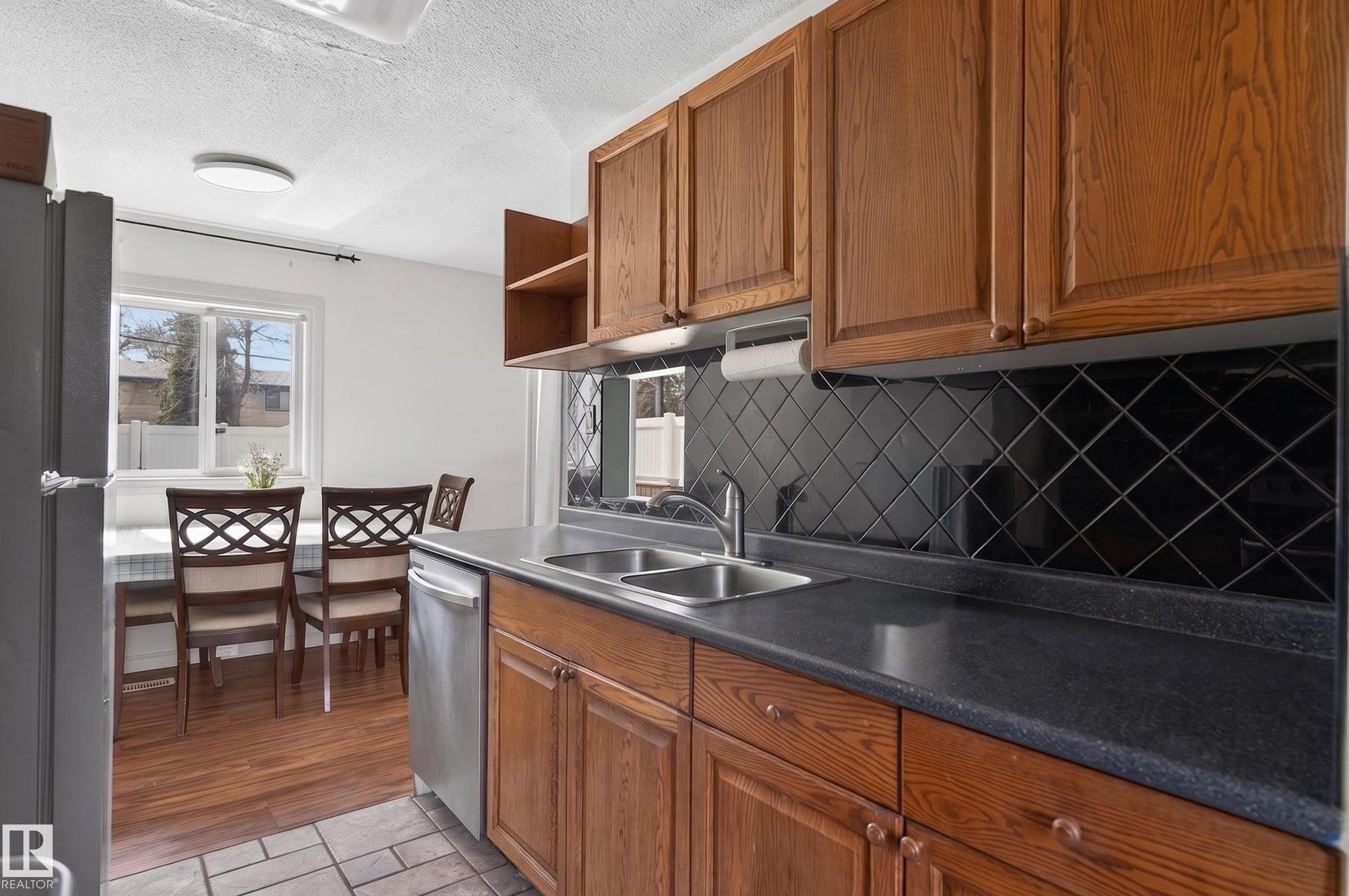 Kitchen featuring warm wood cabinetry, dark countertops, a black diamond-patterned tile backsplash, and a stainless steel double sink - 26E Meadowlark Village, Edmonton, AB - Indoor Photo Showing Kitchen With Double Sink