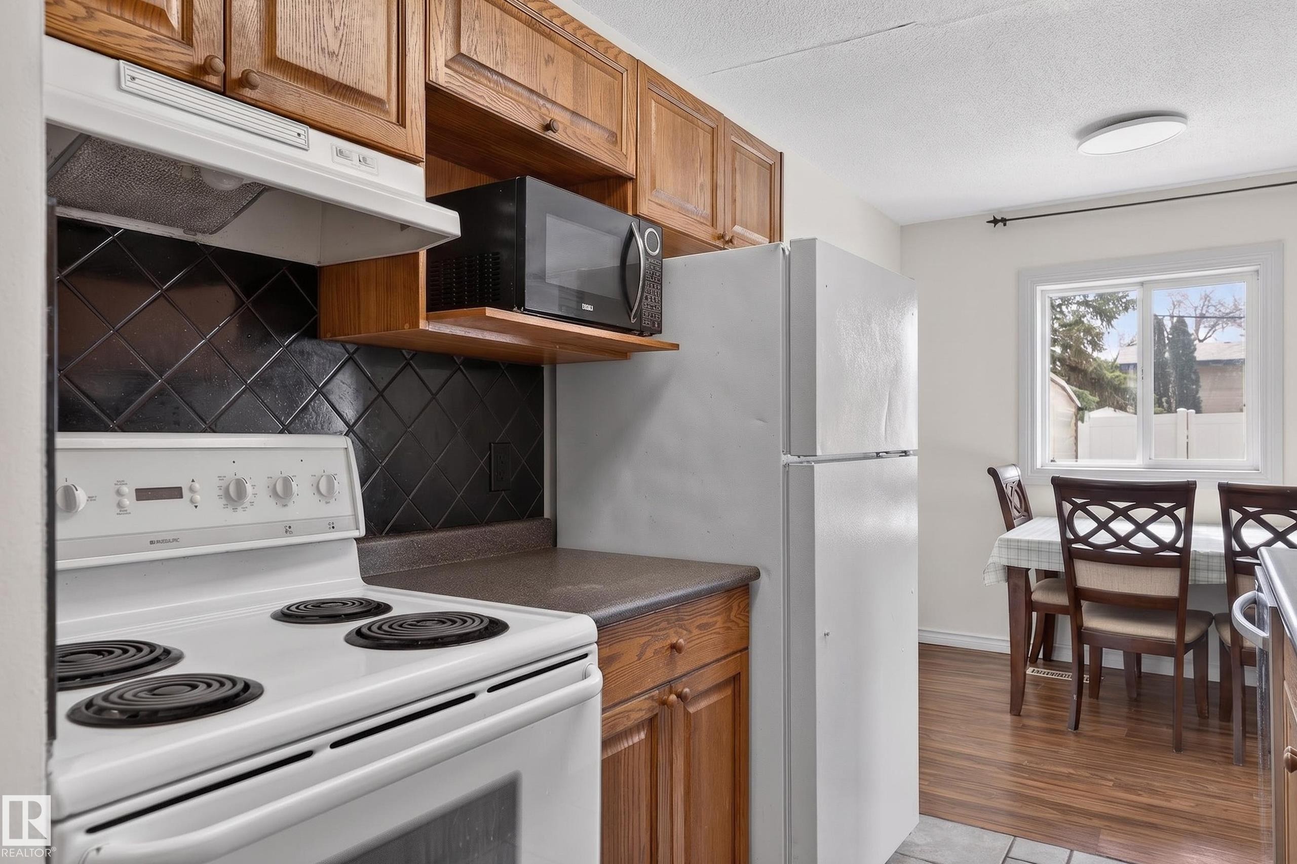 Kitchen featuring wood cabinetry, a white electric range with overhead exhaust, a black diamond-patterned tile backsplash, and a white refrigerator - 26E Meadowlark Village, Edmonton, AB - Indoor Photo Showing Kitchen