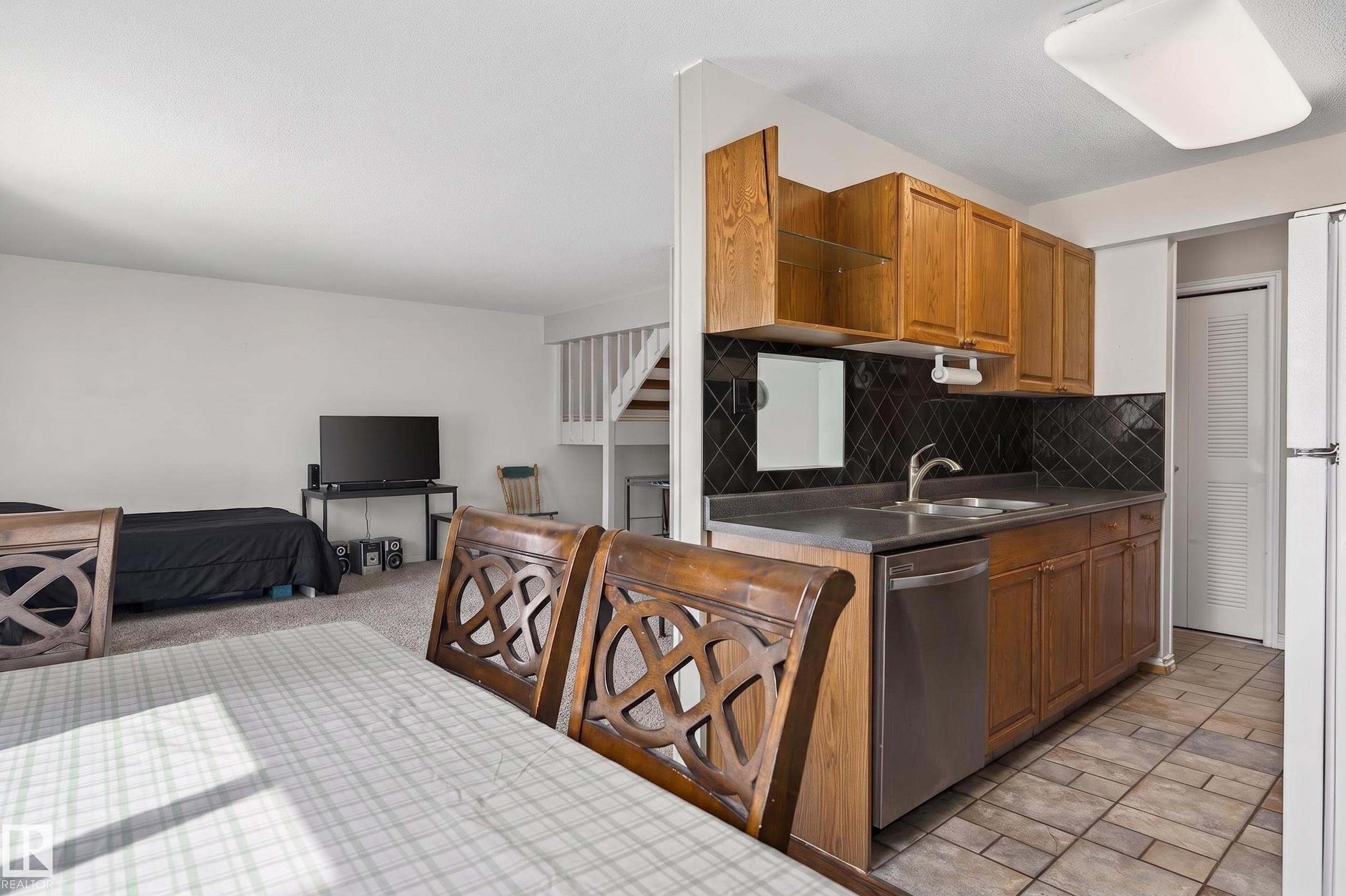 Kitchenette featuring wood cabinetry, stainless steel dishwasher, double basin sink, and dark tile backsplash - 26E Meadowlark Village, Edmonton, AB - Indoor Photo Showing Kitchen With Double Sink