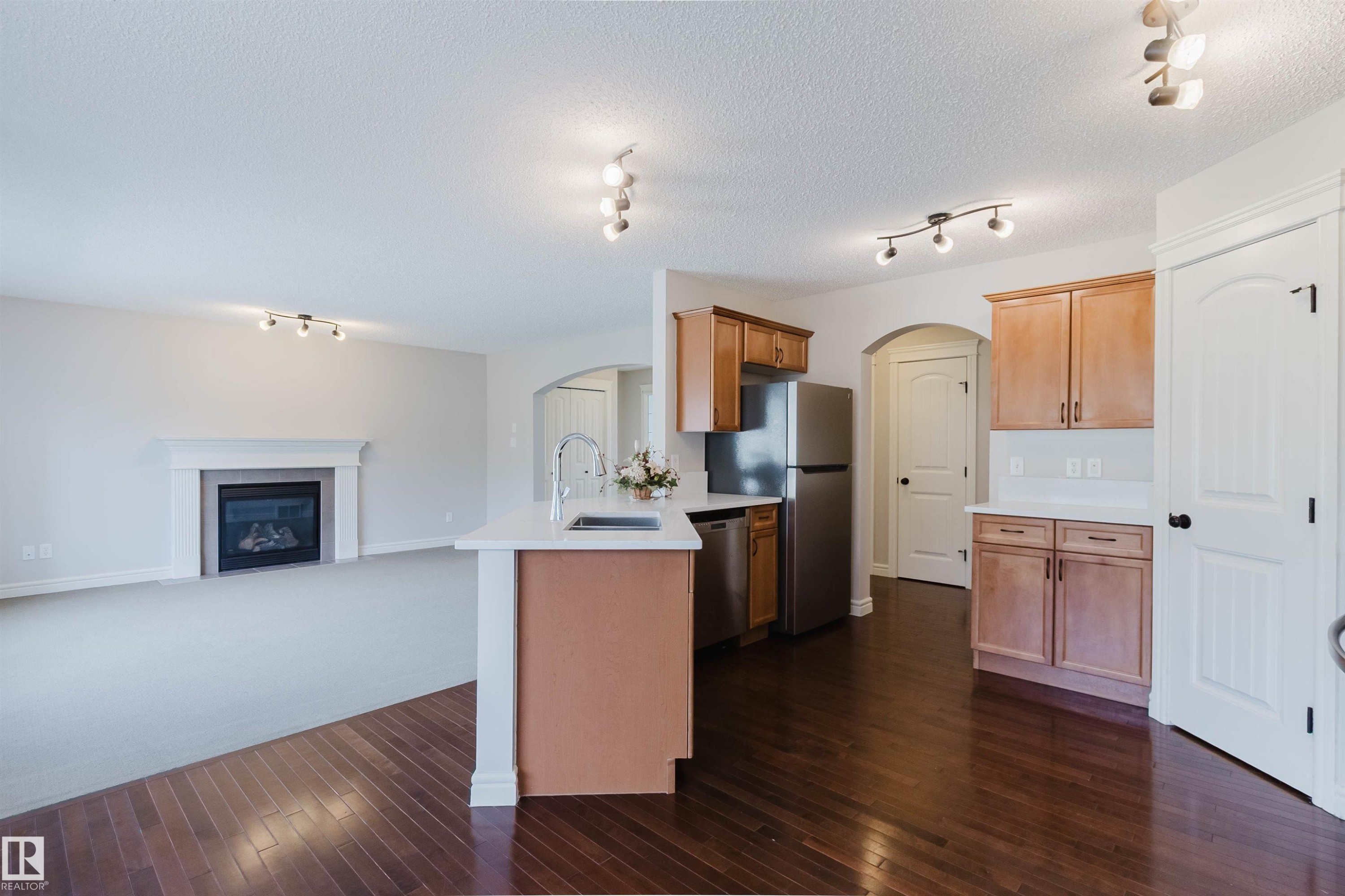 Open-concept kitchen and living area featuring dark wood-finish flooring, a kitchen island with a built-in sink, light wood cabinetry, white countertops, and a living area with a fireplace - 1140 115 Street, Edmonton, AB - Indoor Photo Showing Kitchen With Fireplace