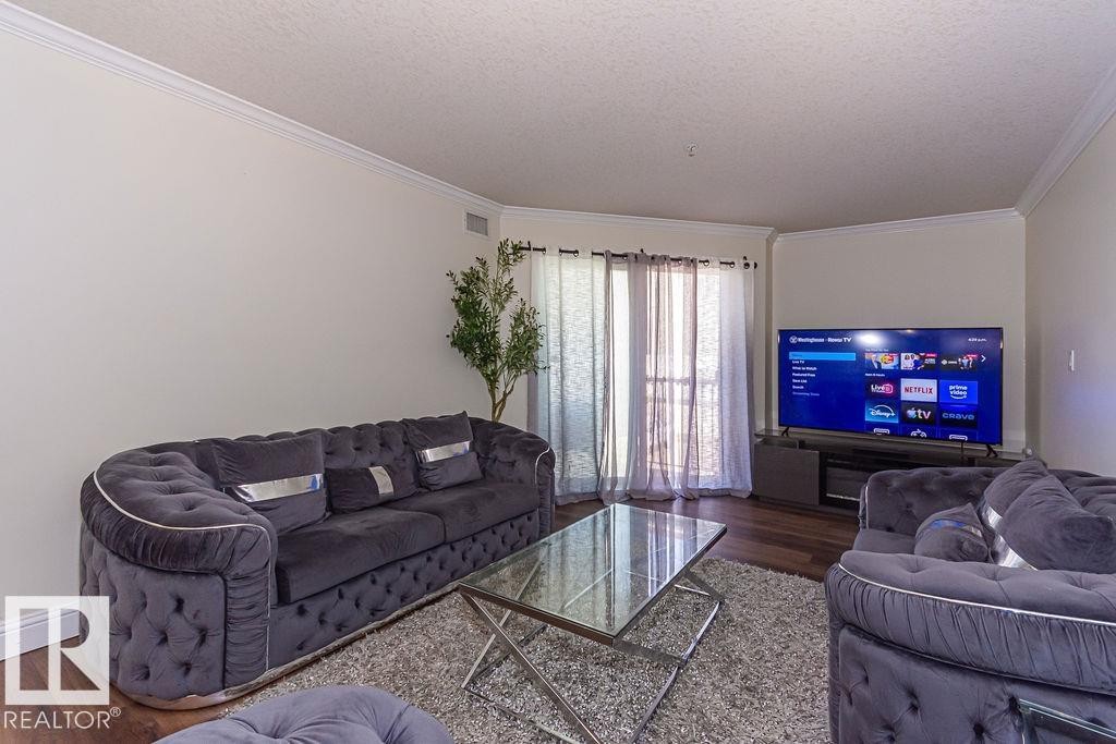 Living area featuring wood-finish flooring, light-toned walls, crown molding, and sliding glass doors with sheer and blackout curtains - 214 13005 140 Avenue, Edmonton, AB - Indoor Photo Showing Living Room