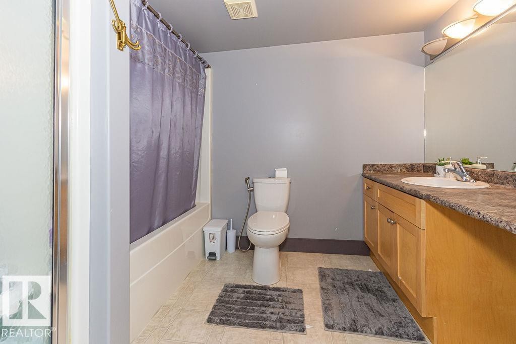 Bathroom featuring a tub/shower combination, light wood-finish vanity with an integrated sink, stone-look countertop, and a wall-mounted mirror - 214 13005 140 Avenue, Edmonton, AB - Indoor Photo Showing Bathroom