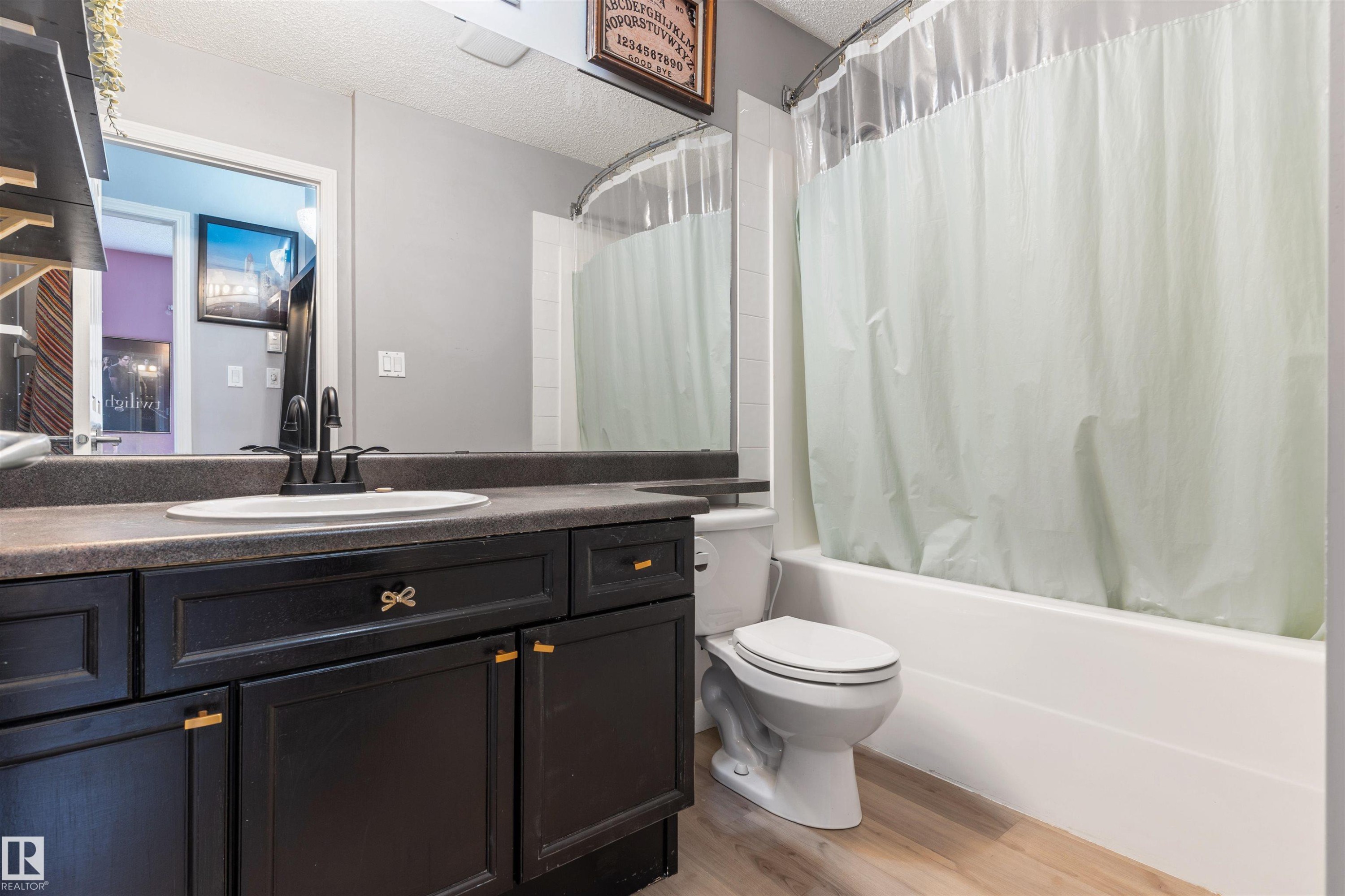 Bathroom featuring a dark wood-finish vanity with a single sink and matte black faucet, a full-width mirror, a white bathtub with shower, and wood-finish flooring - 73 3040 Spence Wynd, Edmonton, AB - Indoor Photo Showing Bathroom