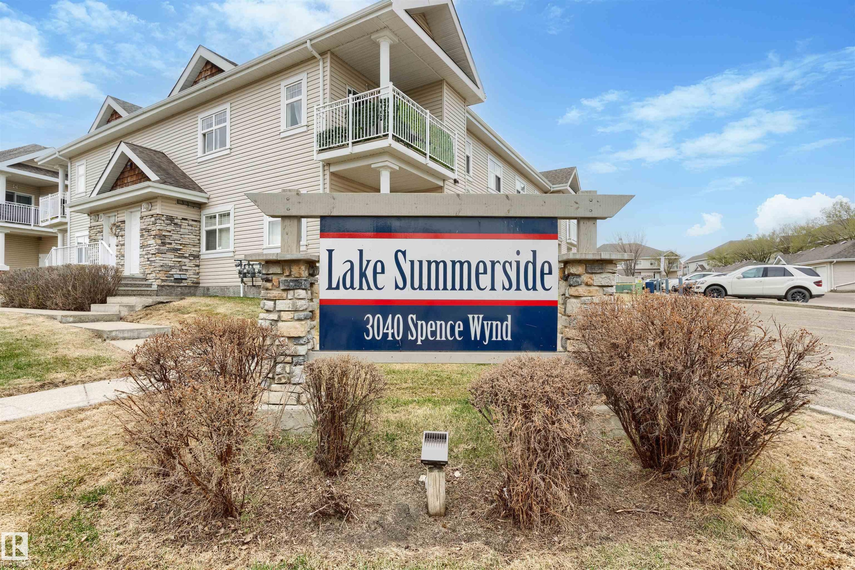 Stone-clad entry sign featuring "Lake Summerside 3040 Spence Wynd" lettering, with a multi-story building exhibiting light-toned siding, white-railed balconies, and stone accents - 73 3040 Spence Wynd, Edmonton, AB - Outdoor