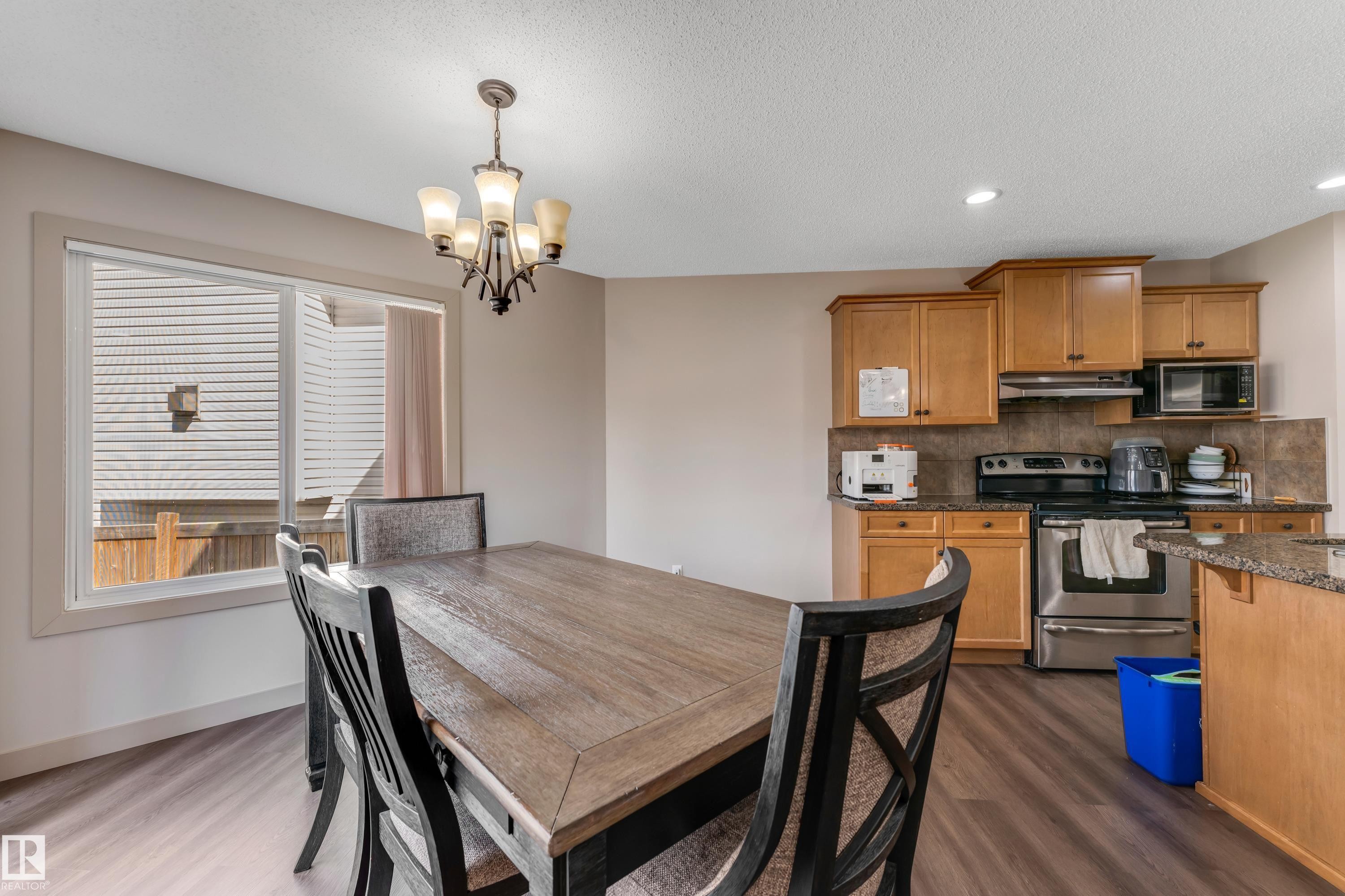 This dining area features a stylish chandelier, ample natural light from the large window, and durable flooring, with a partial view of the kitchen which includes wooden cabinetry, stainless steel appliances, and a tiled backsplash - 463 Tamarack Green, Edmonton, AB - Indoor