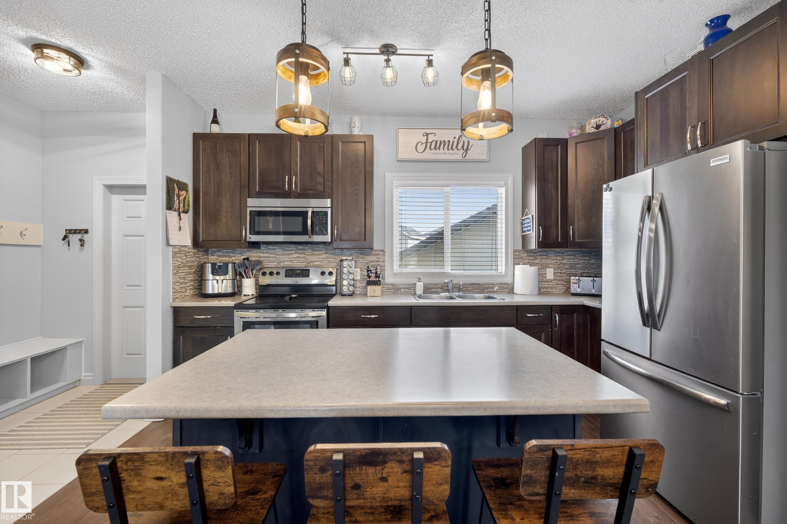 Kitchen featuring a central island with countertop seating, dark wood cabinetry, stainless steel appliances, mosaic tile backsplash, and overhead pendant lighting - 7074 Cardinal Way, Edmonton, AB - Indoor Photo Showing Kitchen With Double Sink