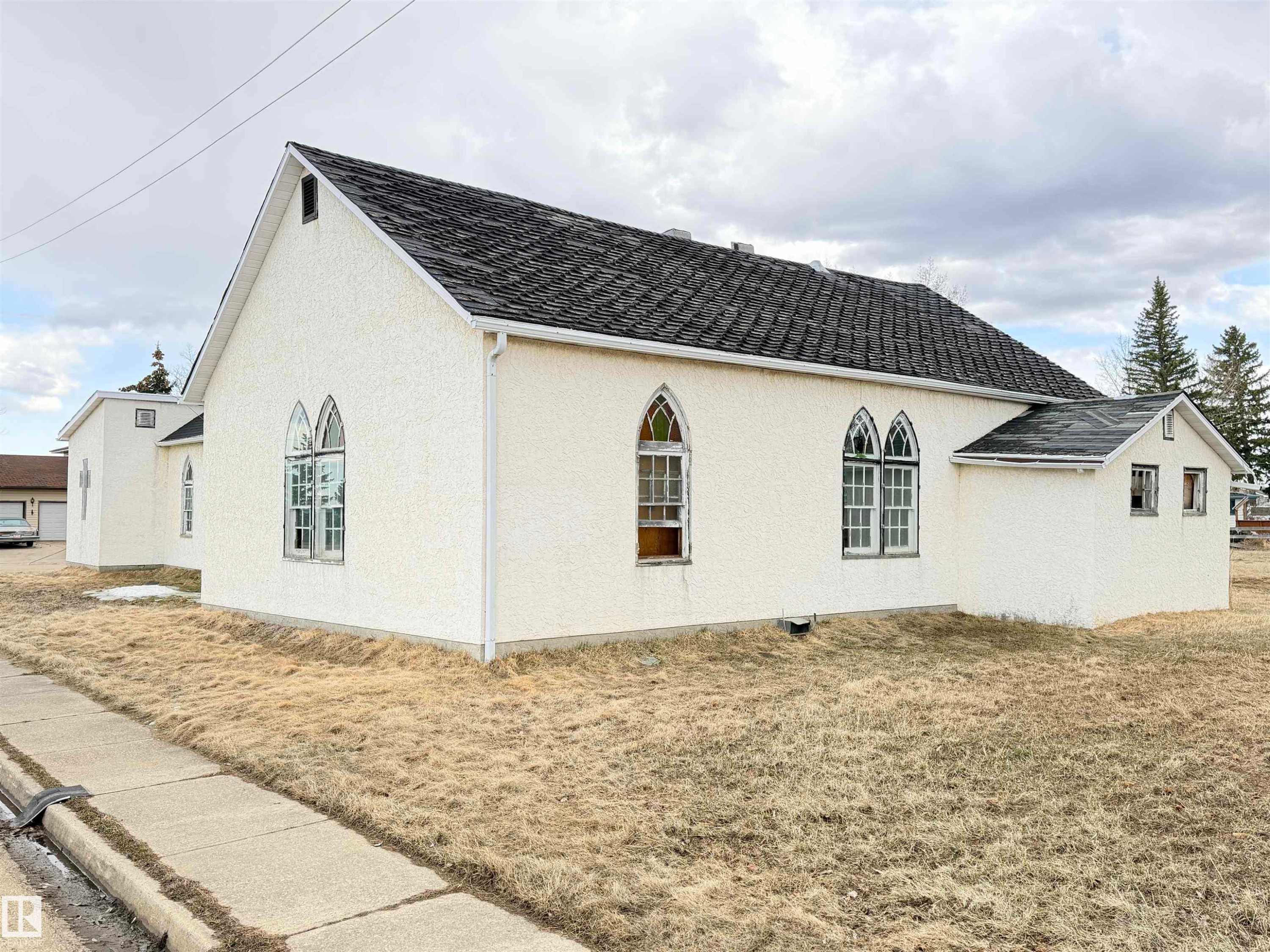 White stucco exterior featuring a gabled roofline with dark shingles, multiple arched windows, and white gutters - 5036 49 Street, Clyde, AB - Outdoor