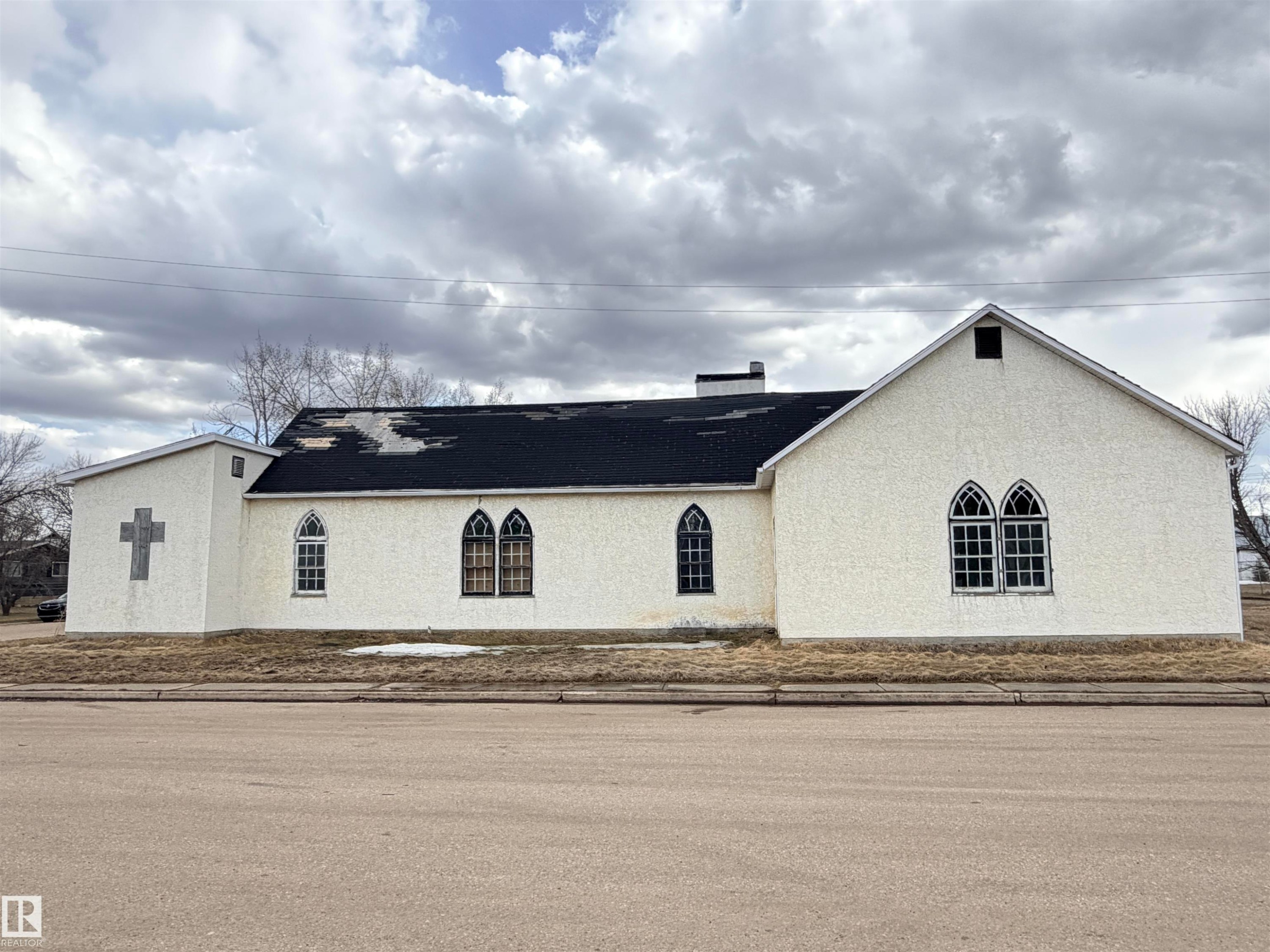White textured stucco exterior with Gothic-style arched windows, a black asphalt shingle roof, and a prominent cross architectural element - 5036 49 Street, Clyde, AB - Outdoor