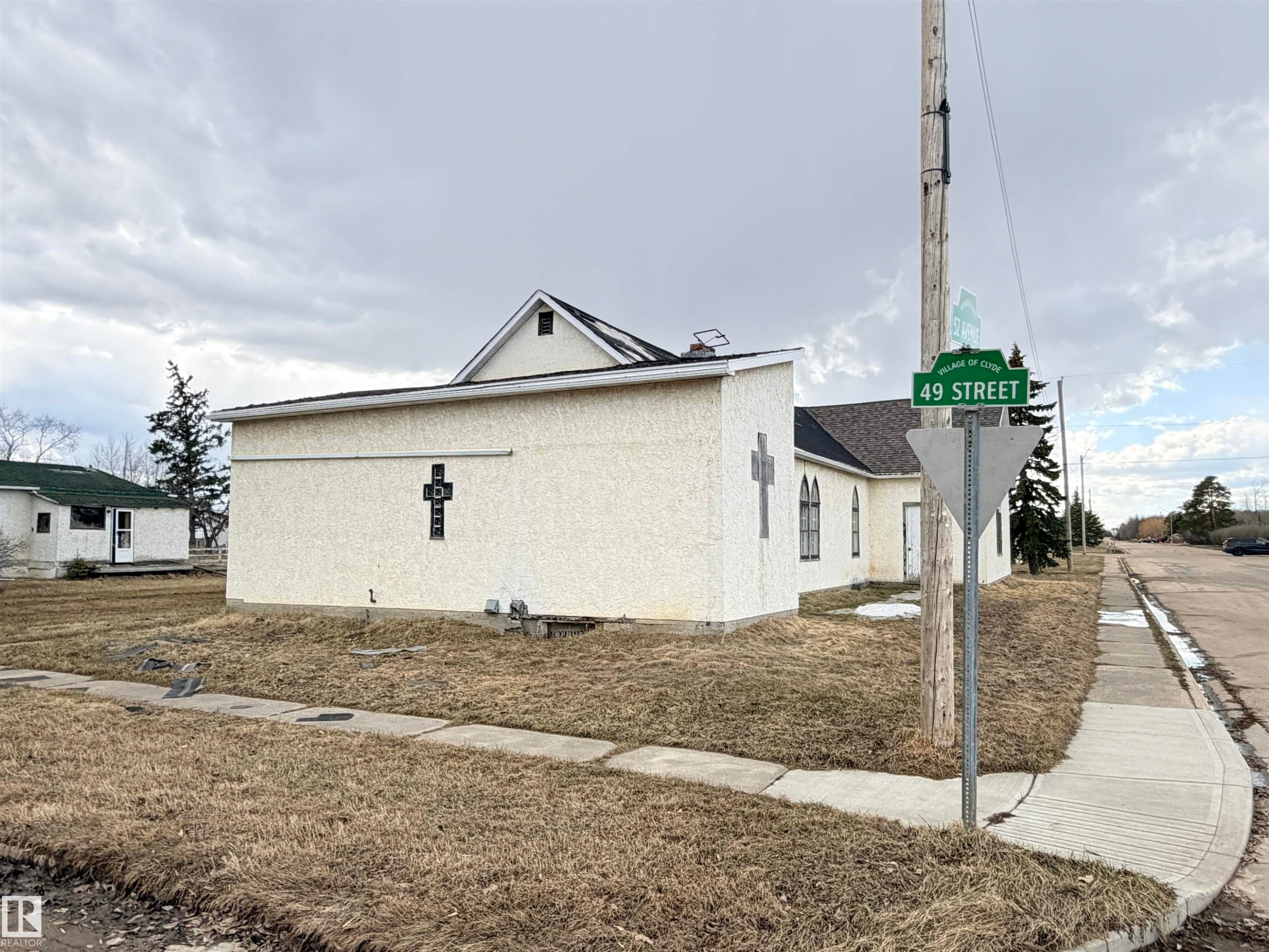 Stucco exterior building with a prominent cross architectural detail, multiple arched windows, and a black asphalt shingle roof - 5036 49 Street, Clyde, AB - Outdoor