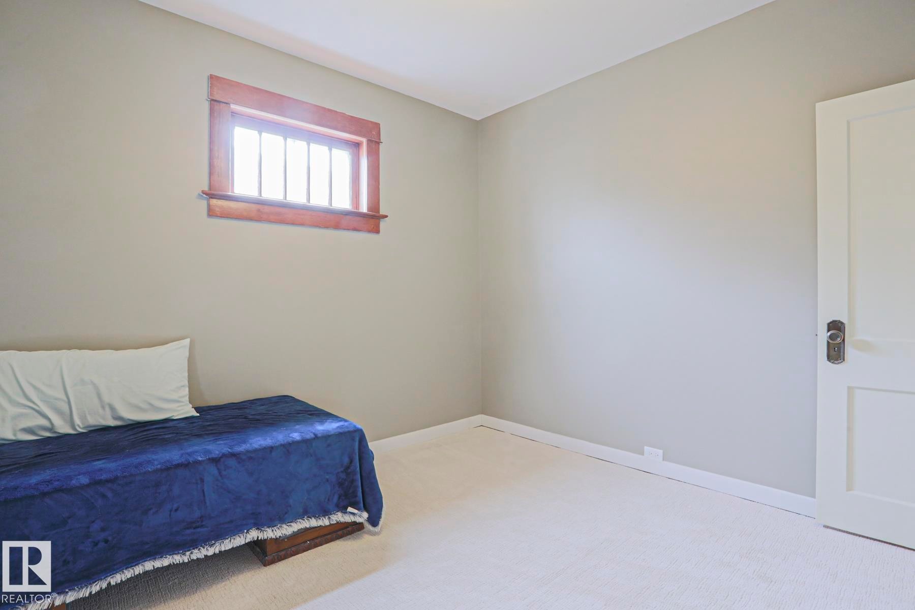 Neutral-toned walls and light-colored carpeting define this room, featuring a window with natural wood trim and multiple vertical muntins - 5113 53 Avenue, Stony Plain, AB - Indoor Photo Showing Bedroom