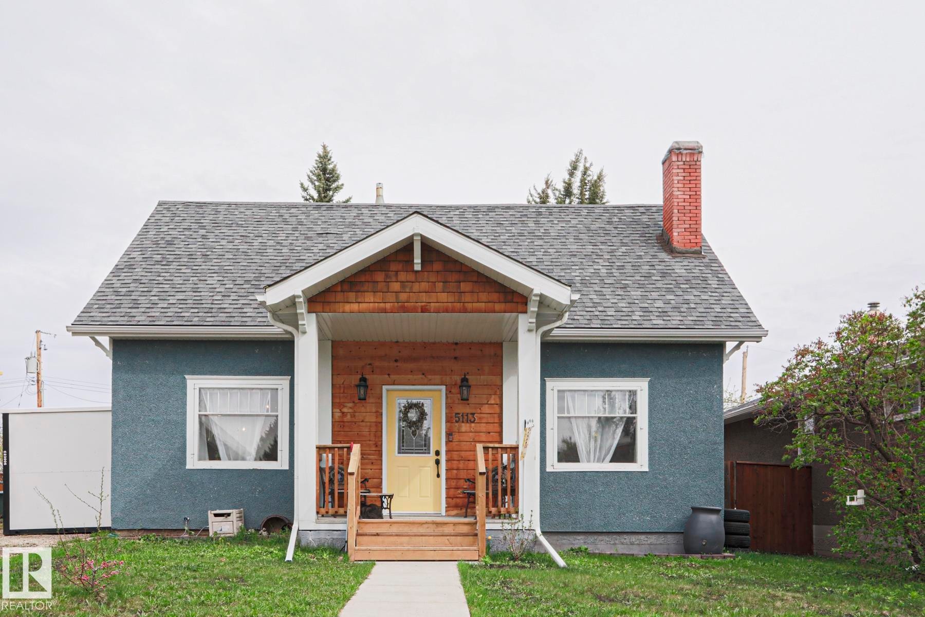 Exterior featuring a blue stucco finish, a covered front porch with wood shingle siding, a yellow front door, a brick chimney, and a gray shingle roof - 5113 53 Avenue, Stony Plain, AB - Outdoor