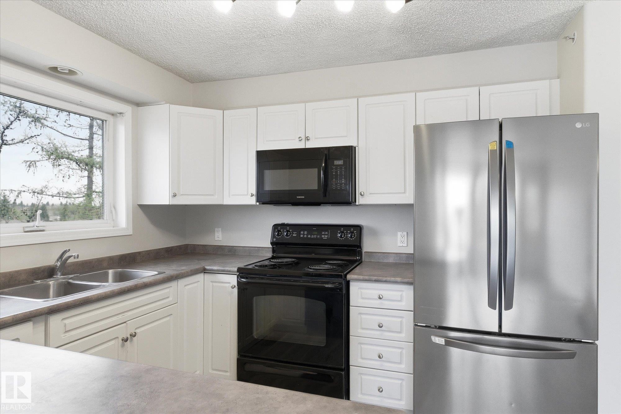 Kitchen featuring white cabinetry, gray countertops, a double basin stainless steel sink, black electric range with an overhead microwave, and a stainless steel French door refrigerator - 407 13635 34 Street Nw, Edmonton, AB - Indoor Photo Showing Kitchen With Double Sink