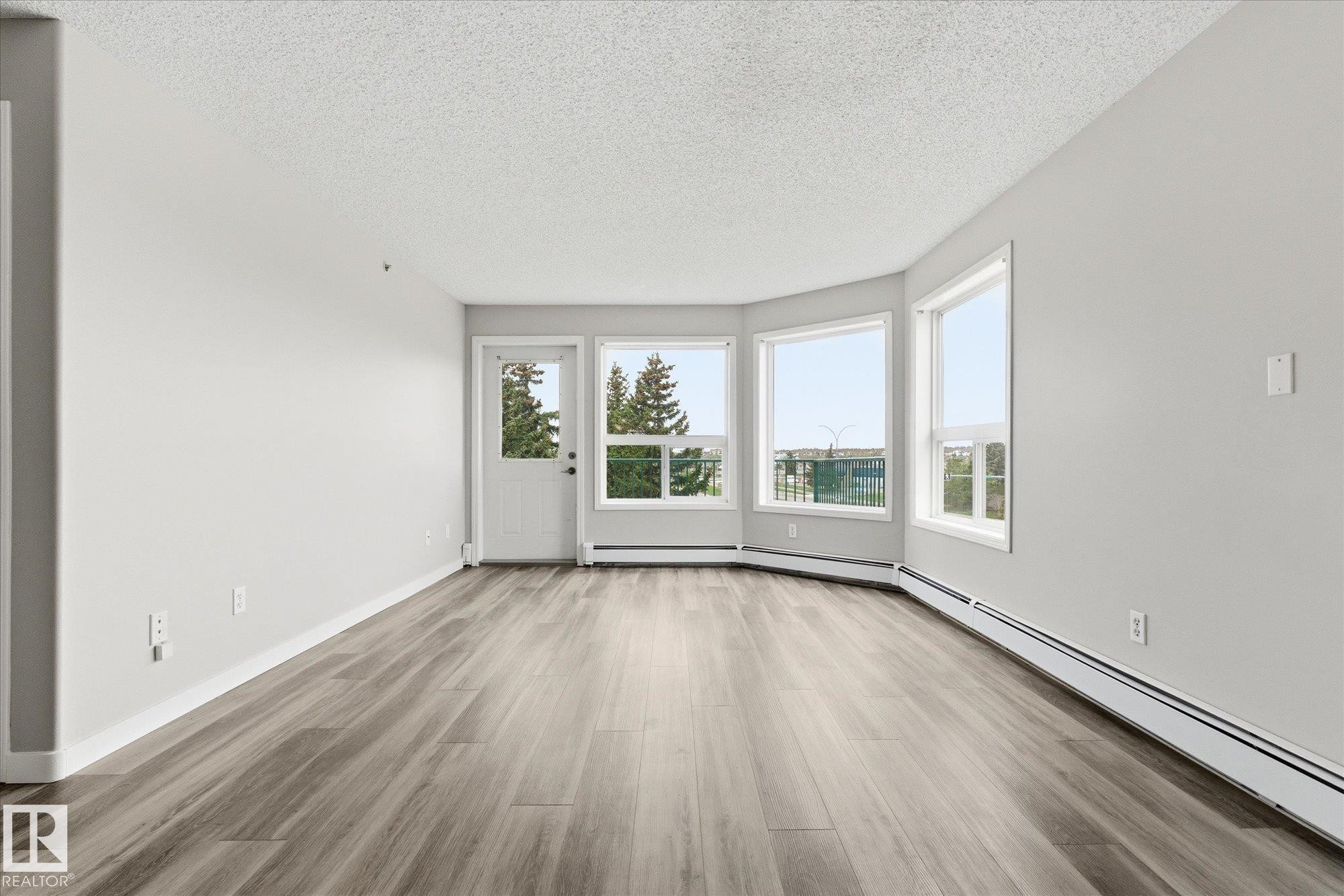 Spacious room featuring wood-finish flooring, light grey walls, and a textured white ceiling - 407 13635 34 Street Nw, Edmonton, AB - Indoor Photo Showing Other Room