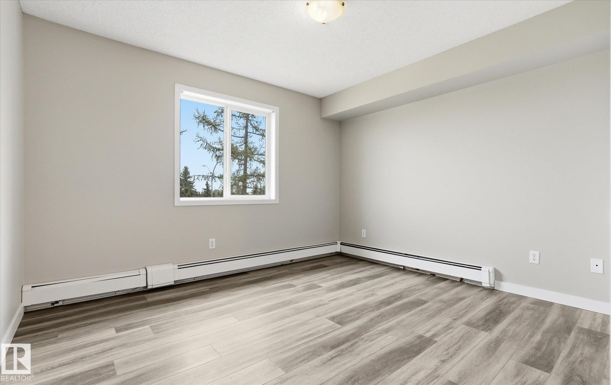 Spacious interior featuring wood-finish flooring, neutral wall tones, a large window, and baseboard heating - 407 13635 34 Street Nw, Edmonton, AB - Indoor Photo Showing Other Room