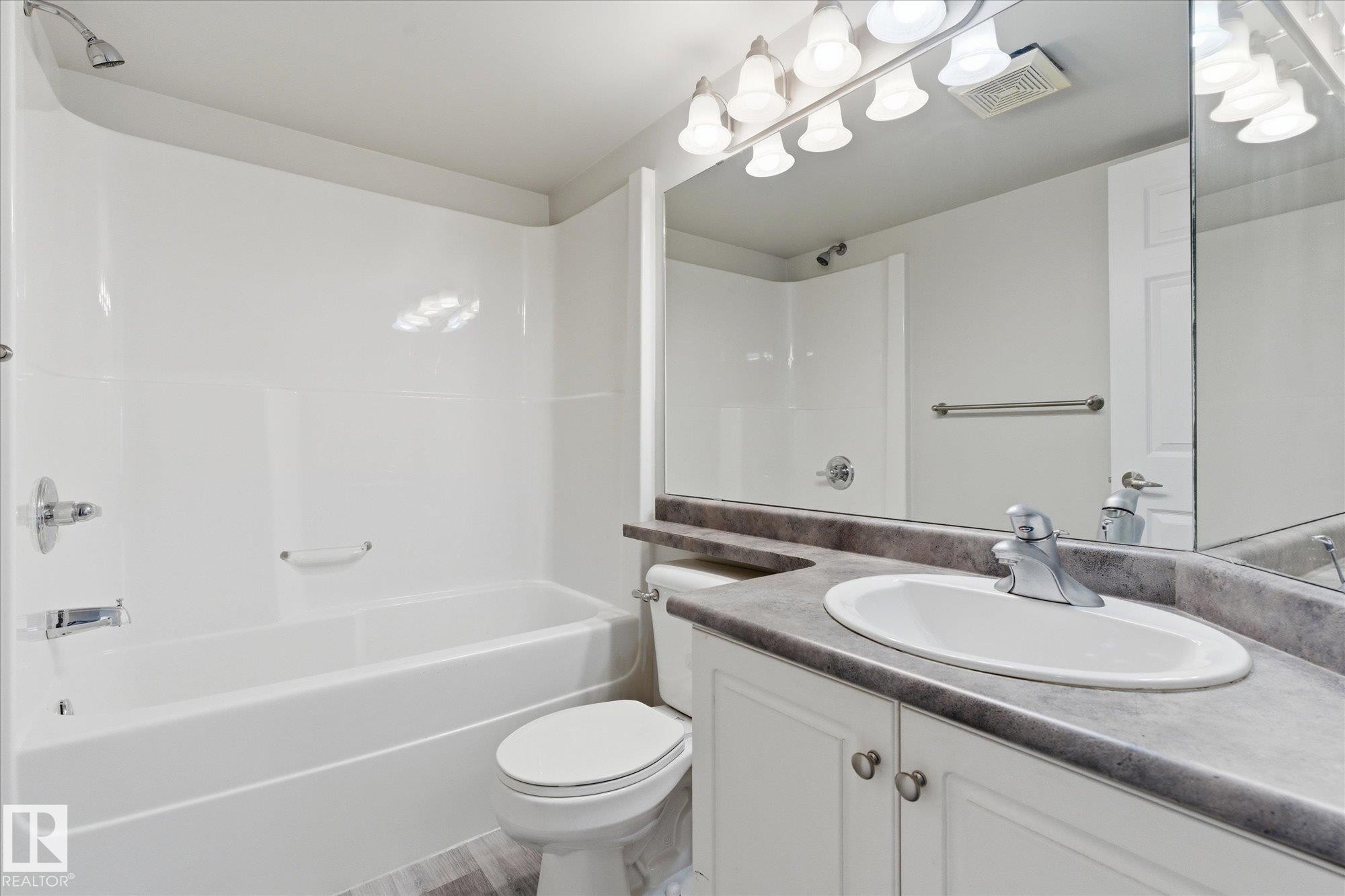 Full bathroom featuring a white tub and shower combination with chrome fixtures, a white toilet, and a vanity with white cabinetry, a gray countertop, and an integrated sink with a chrome faucet - 407 13635 34 Street Nw, Edmonton, AB - Indoor Photo Showing Bathroom