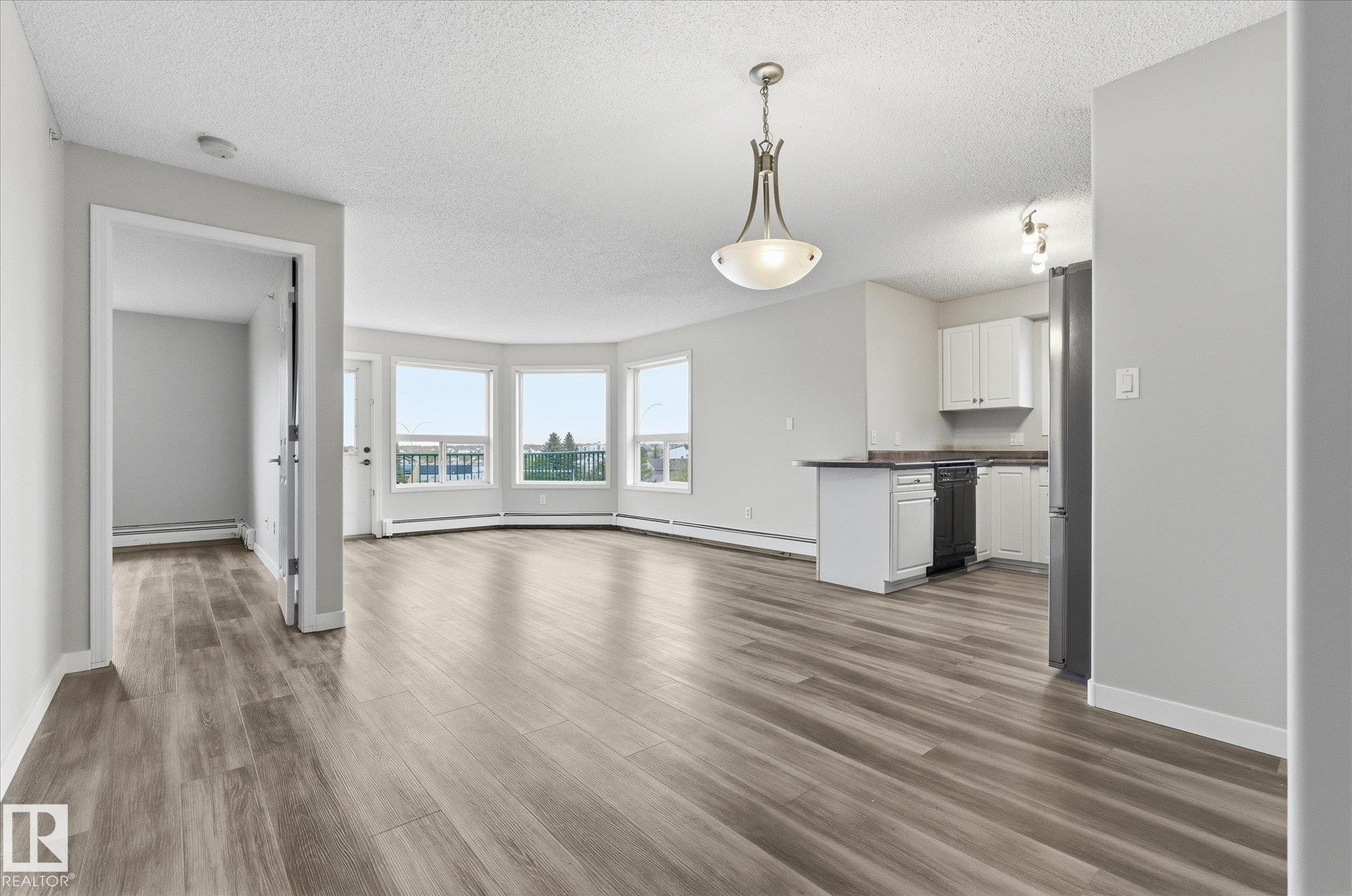 Open-concept living area with wood-finish flooring, a bay window offering exterior scenery, and a kitchen featuring white cabinetry and black appliances - 407 13635 34 Street Nw, Edmonton, AB - Indoor Photo Showing Other Room