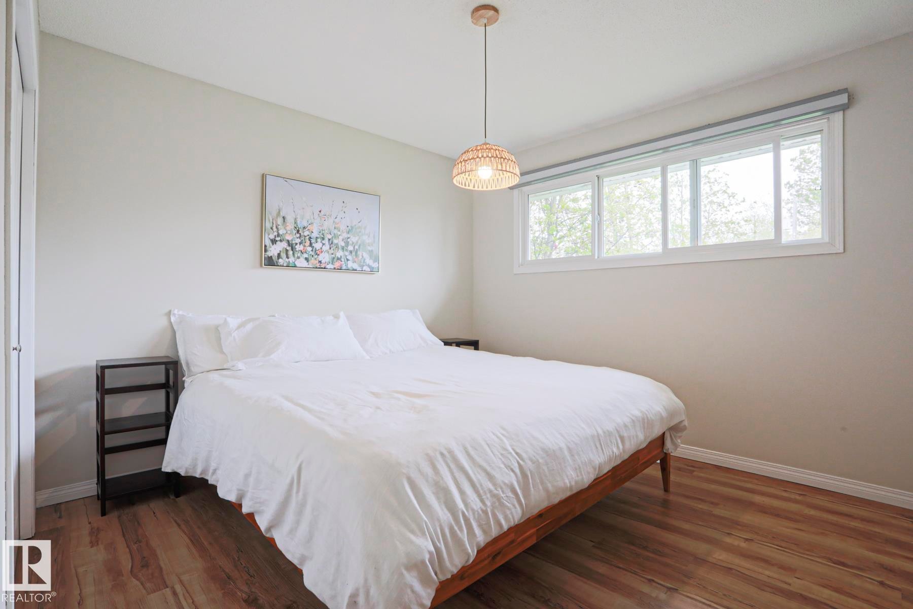 Bright room featuring wood-finish flooring, a large window with roller shades, and a woven pendant light fixture - 9523 151 Street, Edmonton, AB - Indoor Photo Showing Bedroom