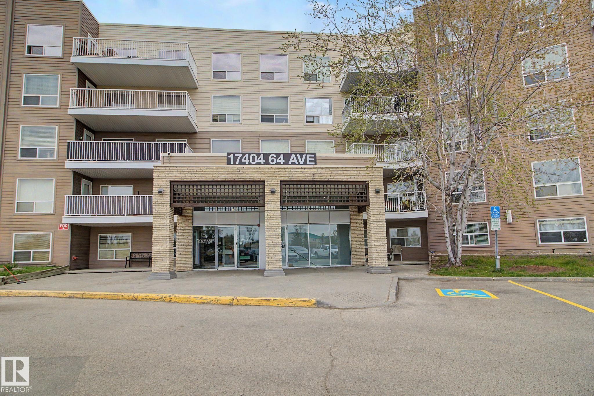 Multi-story residential building featuring a prominent stone facade entrance with a metal canopy, multiple private balconies with white railings, and a dedicated accessible parking space - 213 17404 64 Avenue, Edmonton, AB