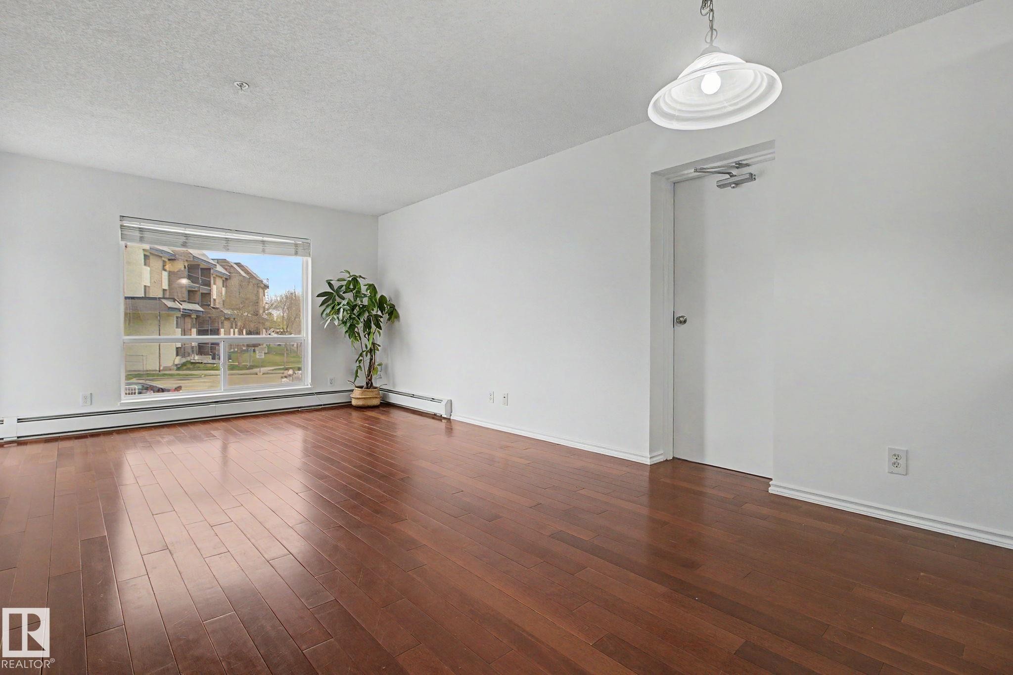 Spacious interior featuring wood-finish flooring, a large window, white walls, and a ceiling-mounted light fixture - 213 17404 64 Avenue, Edmonton, AB