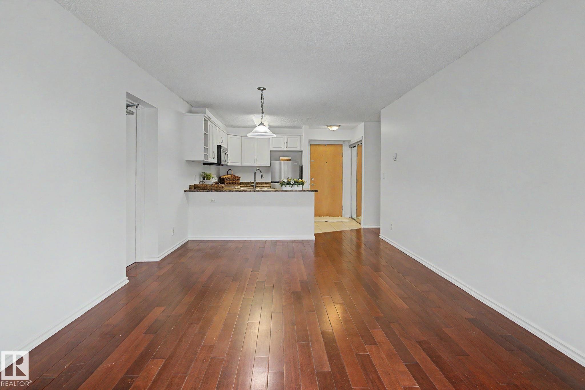 Hardwood flooring throughout a bright living area, featuring a kitchen with white cabinetry, light-toned countertops, and a stainless steel refrigerator - 213 17404 64 Avenue, Edmonton, AB