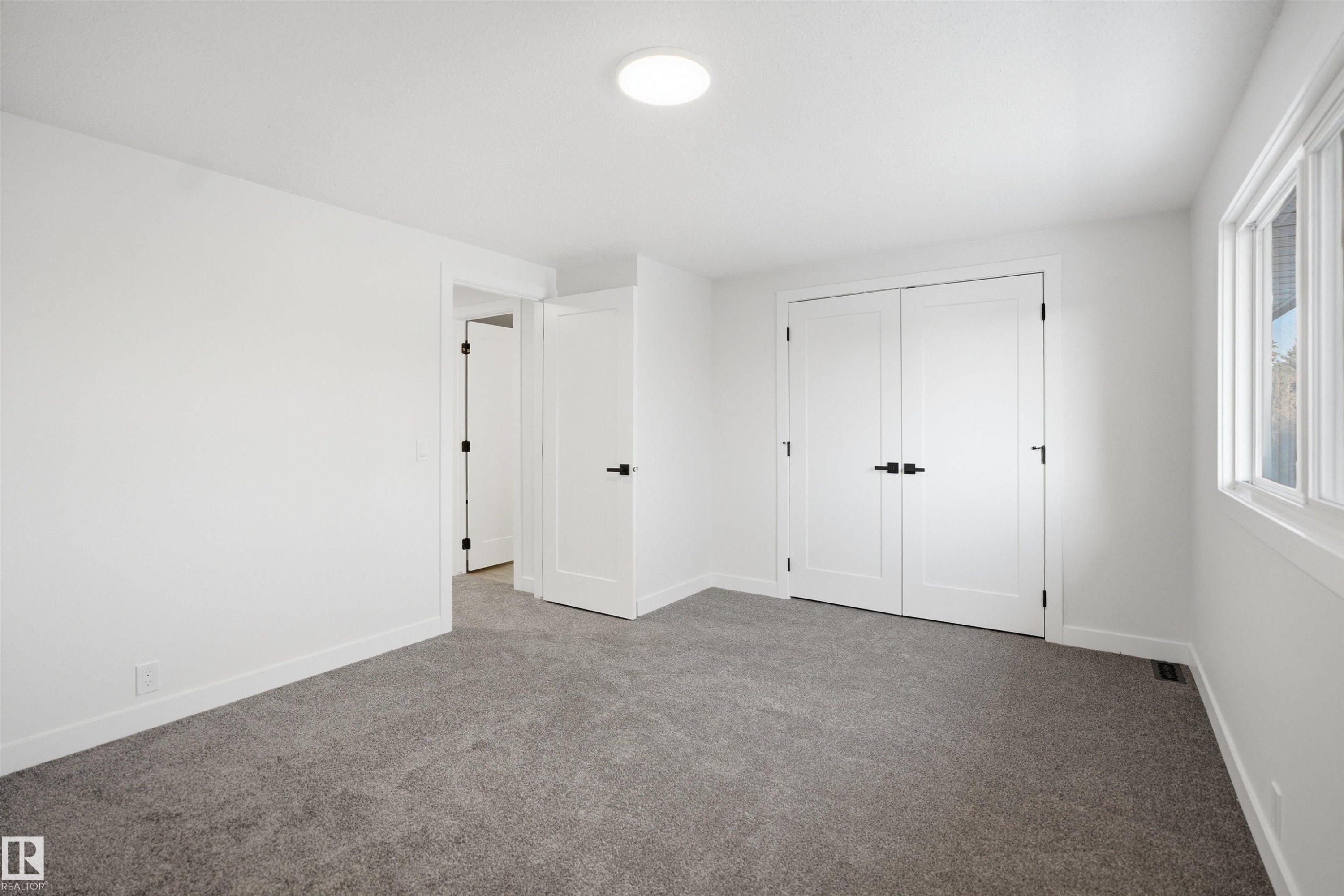 Spacious room featuring neutral carpeting, white walls, a large window with white trim, double panel doors with black hardware, and a circular flush-mount ceiling light - 1706 37 Street, Edmonton, AB - Indoor Photo Showing Other Room