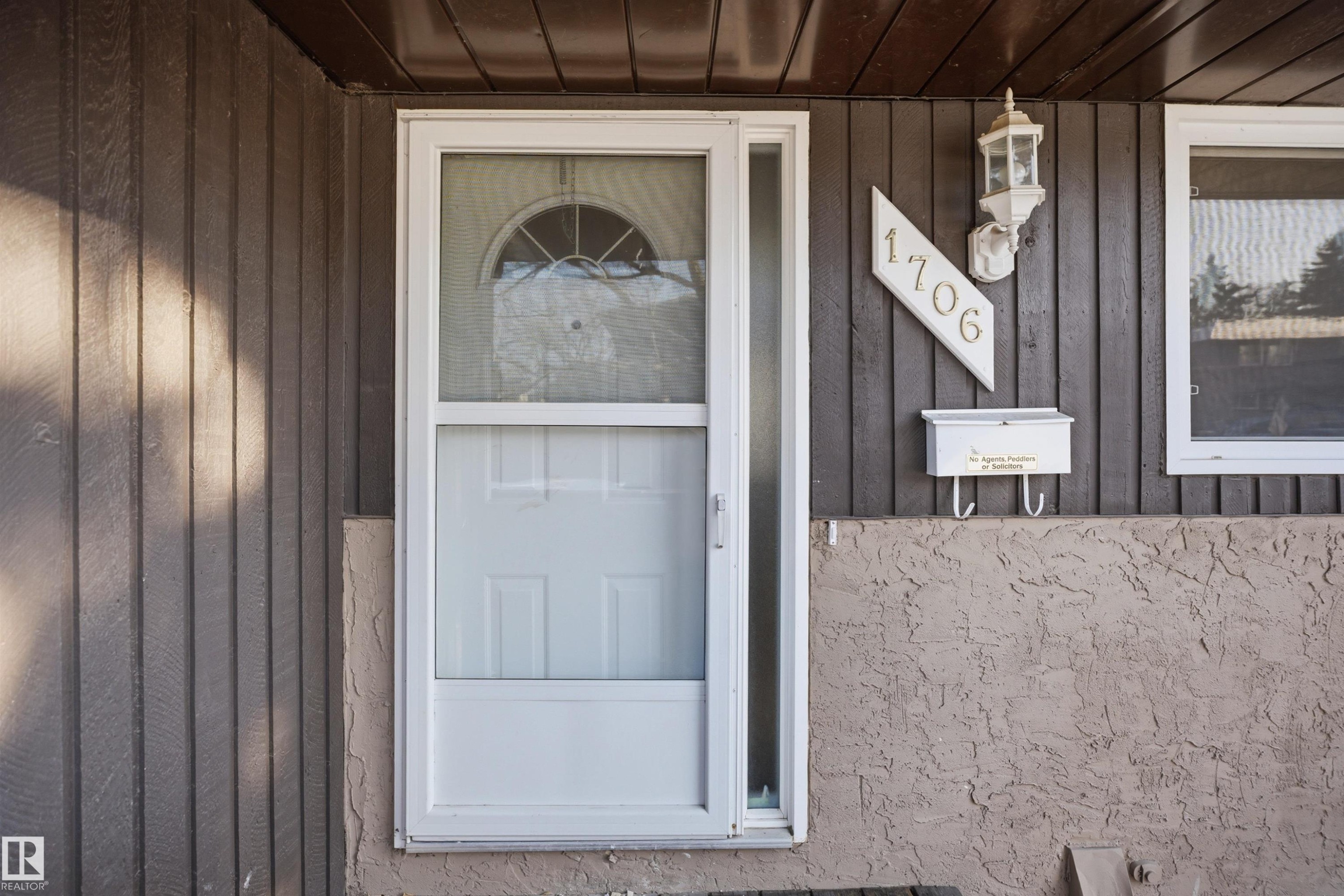 Front entry with a white storm door, vertical wood panel siding, decorative outdoor lighting, and a textured stucco foundation - 1706 37 Street, Edmonton, AB - Outdoor With Exterior