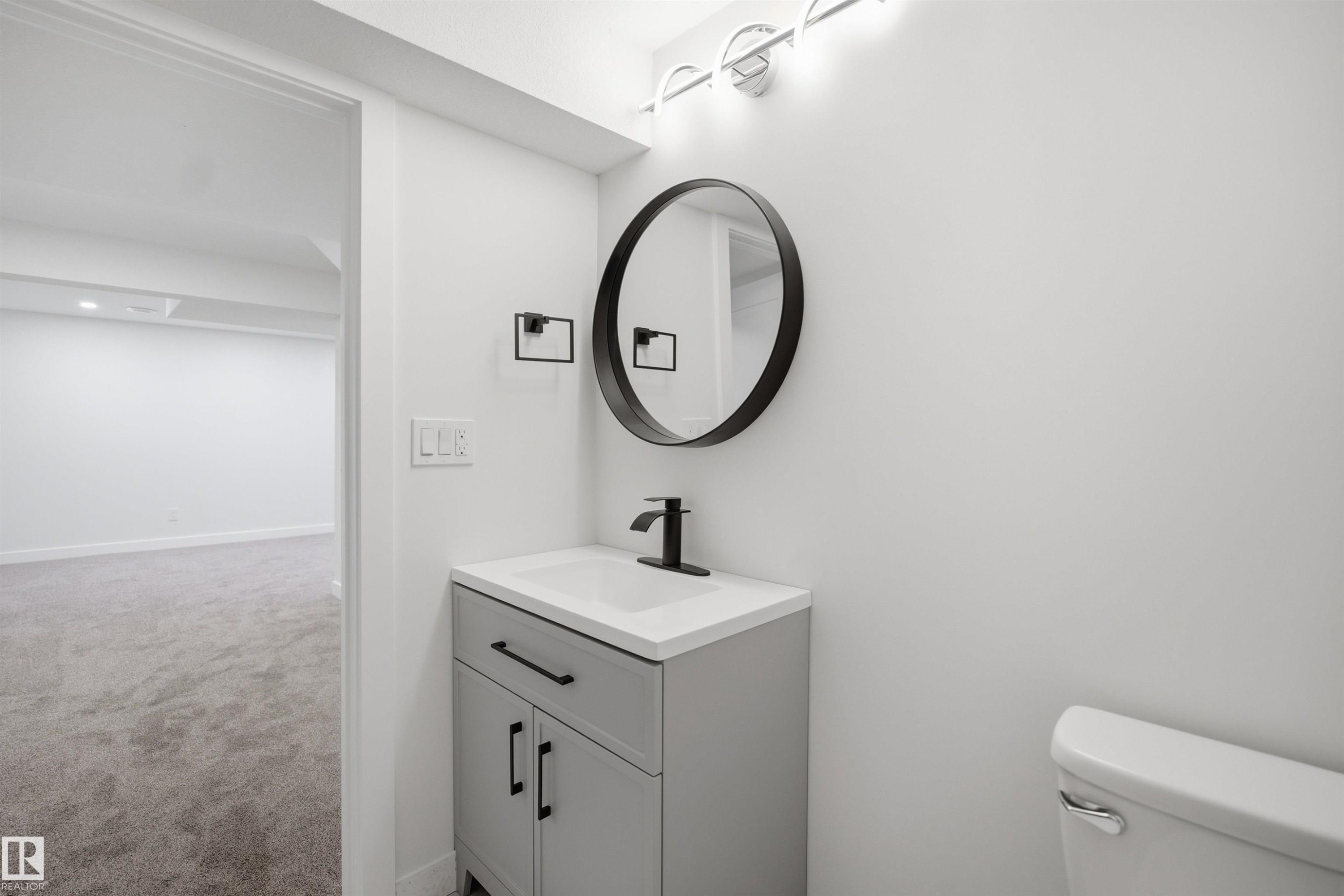 Contemporary bathroom featuring a vanity with a white countertop and matte black hardware, a round framed mirror, and modern wall-mounted lighting - 1706 37 Street, Edmonton, AB - Indoor Photo Showing Bathroom