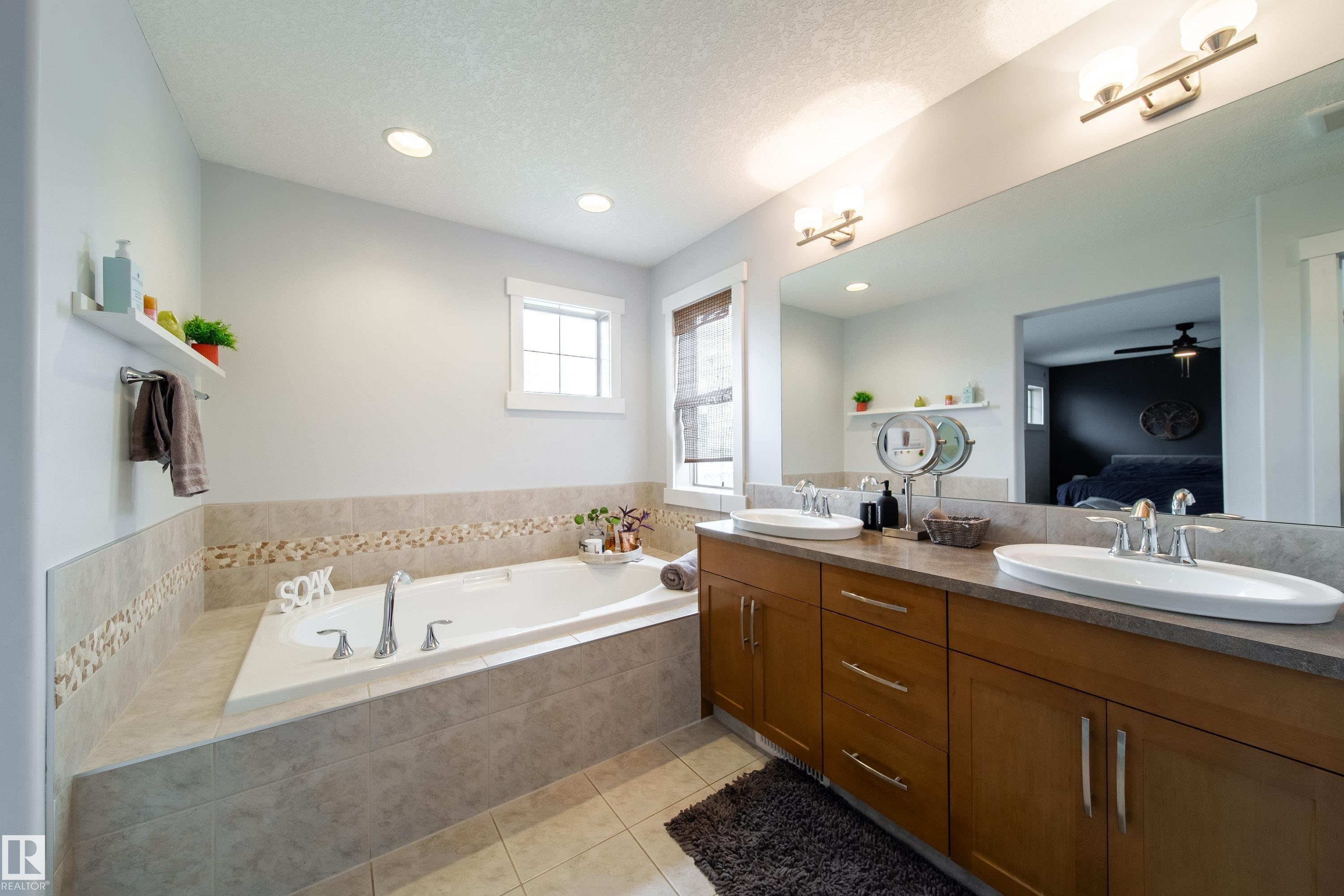 Spacious bathroom featuring a built-in soaking tub with tiled surround, a double vanity with wood-finish cabinetry, dual undermount sinks, and a large wall-mounted mirror - 2016 125 Street, Edmonton, AB - Indoor Photo Showing Bathroom
