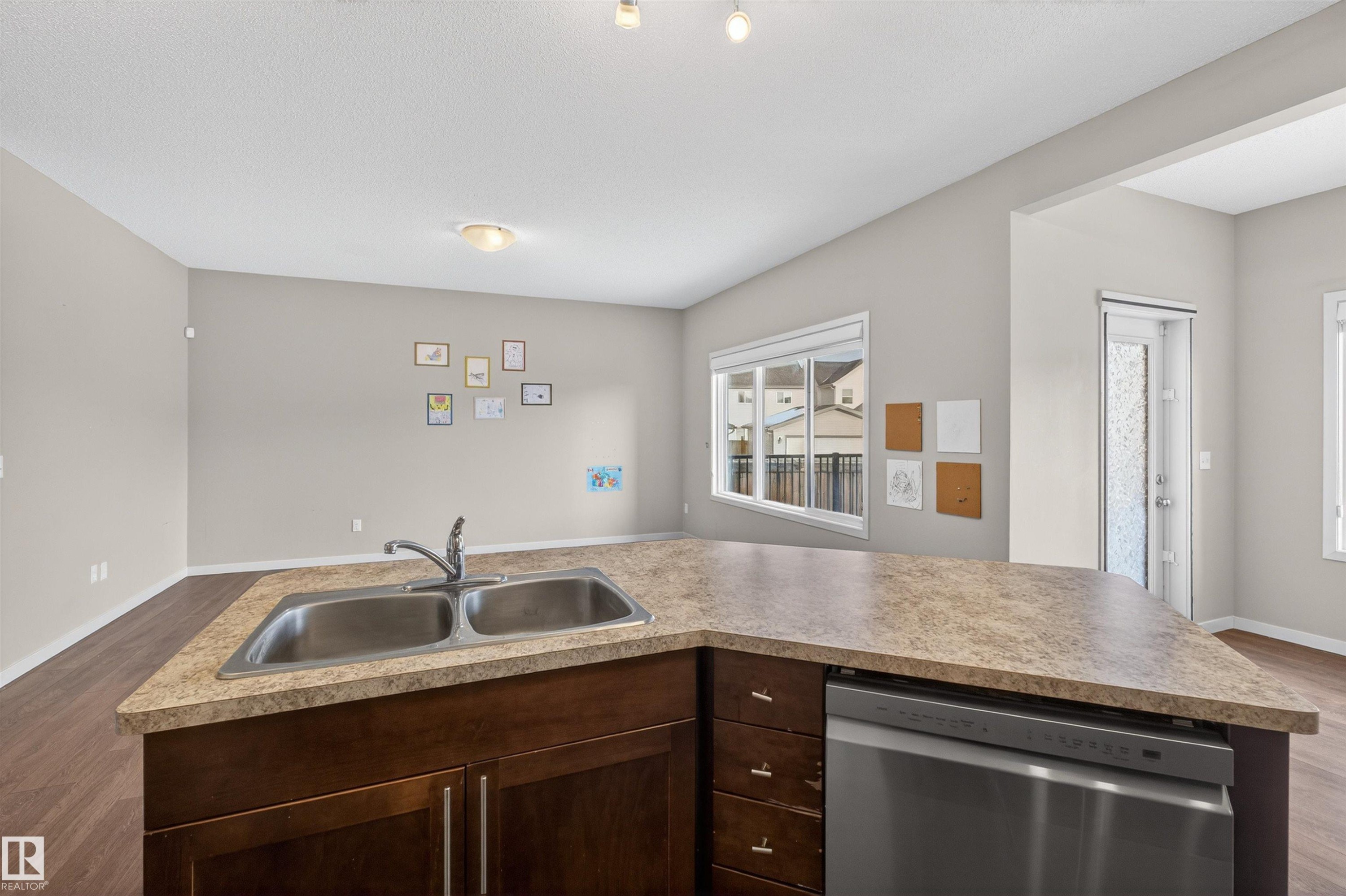 Kitchen island featuring a dual-basin stainless steel sink with a single-lever faucet, rich wood-finish cabinetry, and a built-in stainless steel dishwasher - 4723 Alwood Bend Sw, Edmonton, AB - Indoor Photo Showing Kitchen With Double Sink
