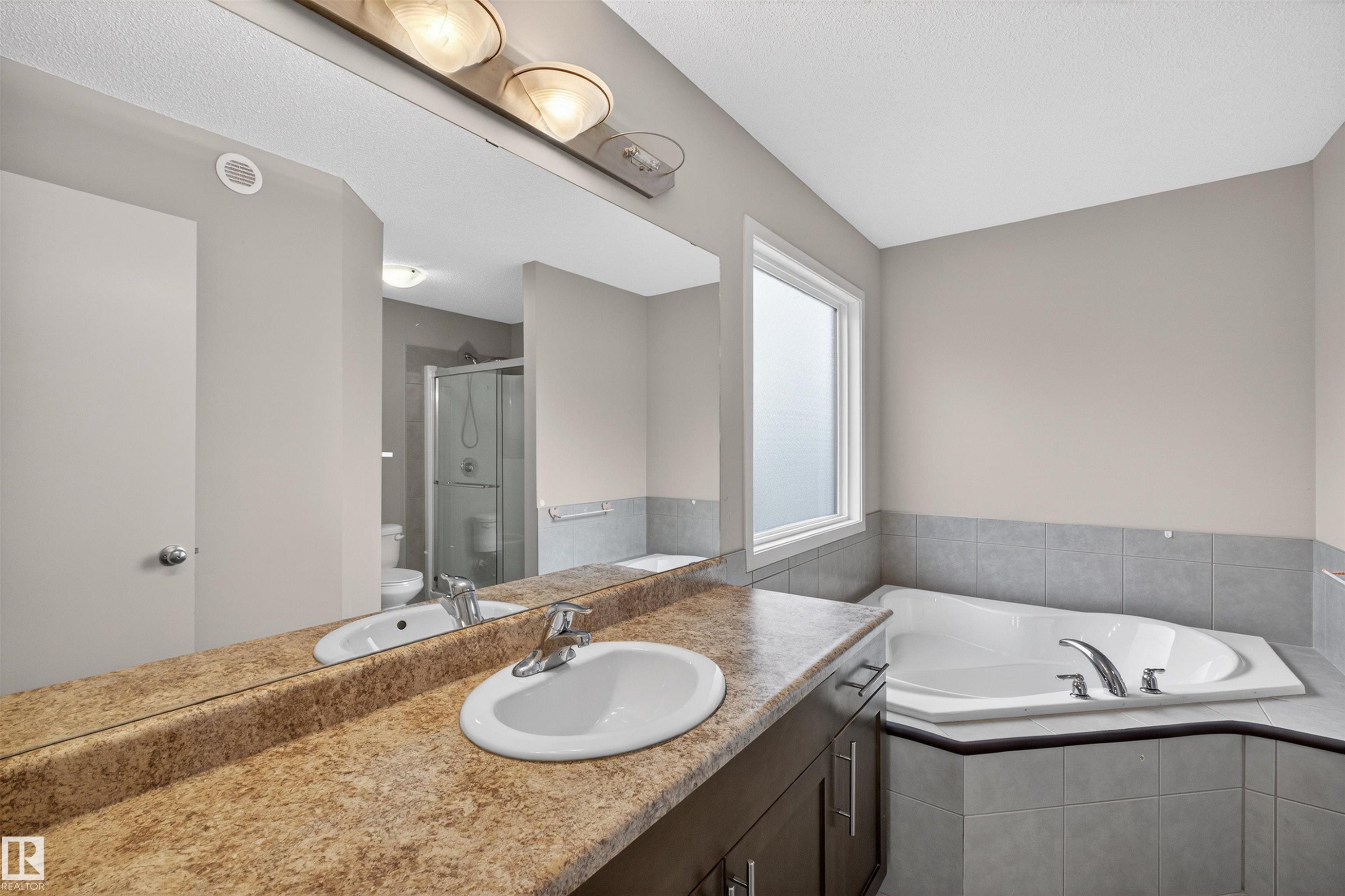 Bathroom featuring an integrated vanity with a single basin sink, an expansive wall mirror, an adjacent window-lit corner soaking tub, and a separate glass-enclosed shower - 4723 Alwood Bend Sw, Edmonton, AB - Indoor Photo Showing Bathroom