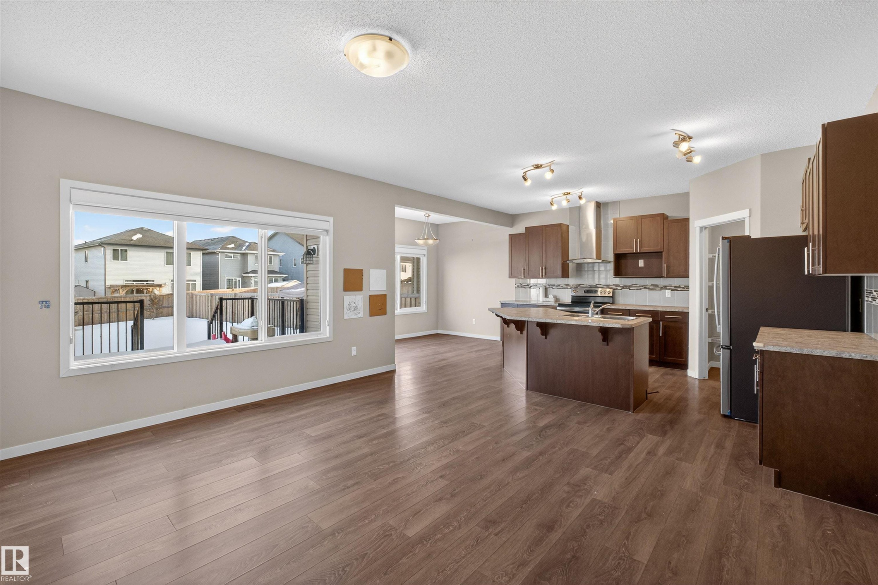 Open-concept living space featuring wood-finish flooring, a contemporary kitchen island with seating, dark wood cabinetry, and stainless steel appliances - 4723 Alwood Bend Sw, Edmonton, AB - Indoor Photo Showing Kitchen