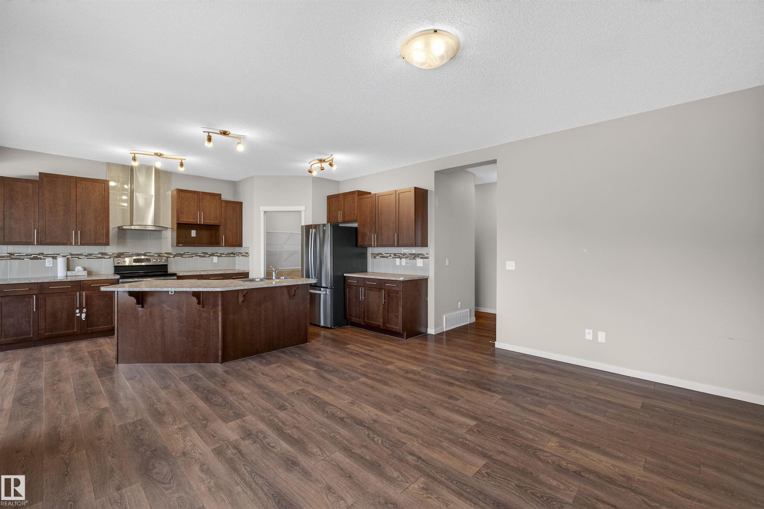 Kitchen featuring wood-finish flooring, dark wood cabinetry, a central island with integrated sink, stainless steel appliances, and a tiled backsplash - 4723 Alwood Bend Sw, Edmonton, AB - Indoor Photo Showing Kitchen