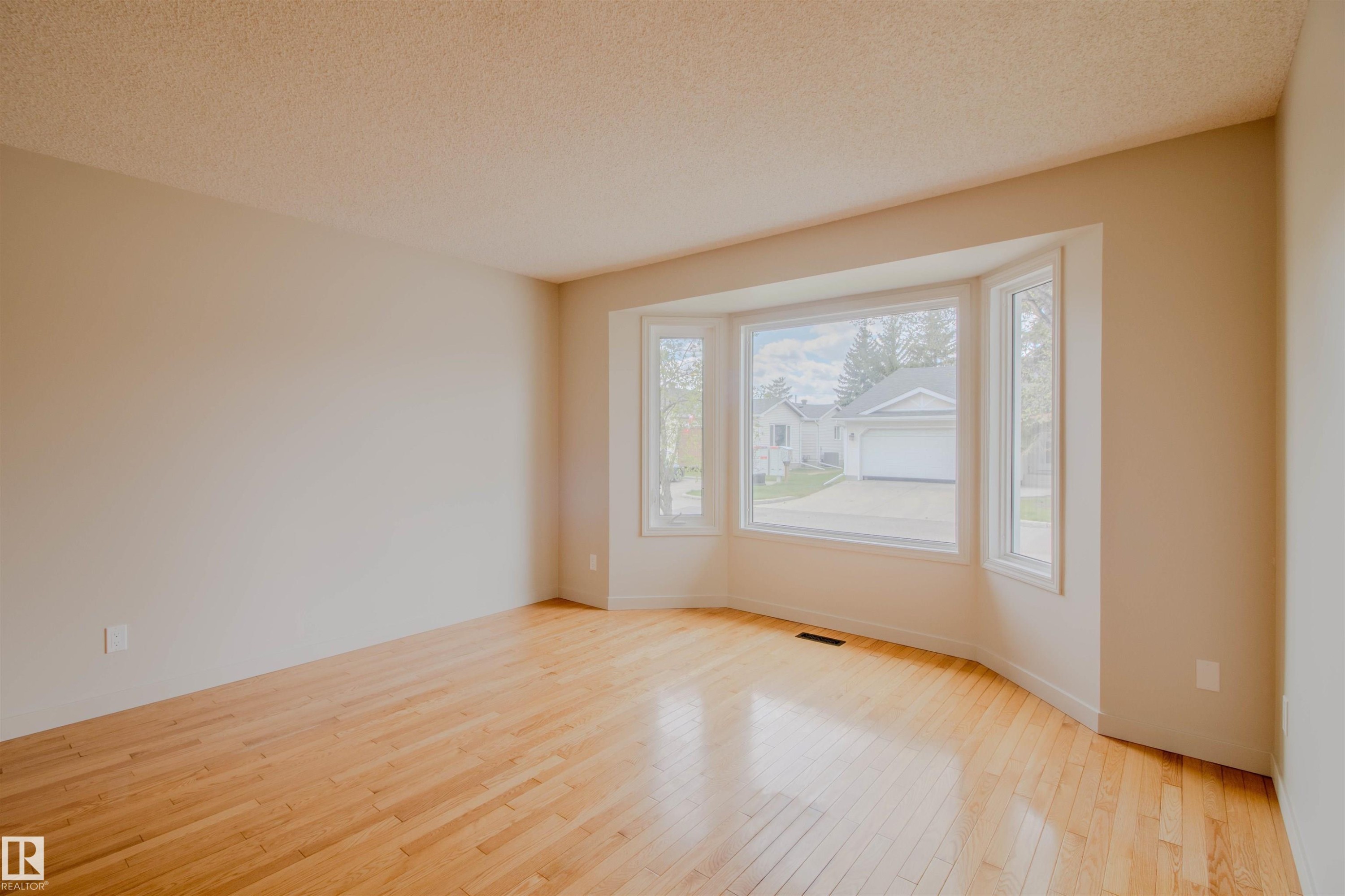 Spacious room featuring a prominent bay window, light wood-finish flooring, neutral wall paint, and a textured ceiling - 5935 189 Street, Edmonton, AB - Indoor Photo Showing Other Room
