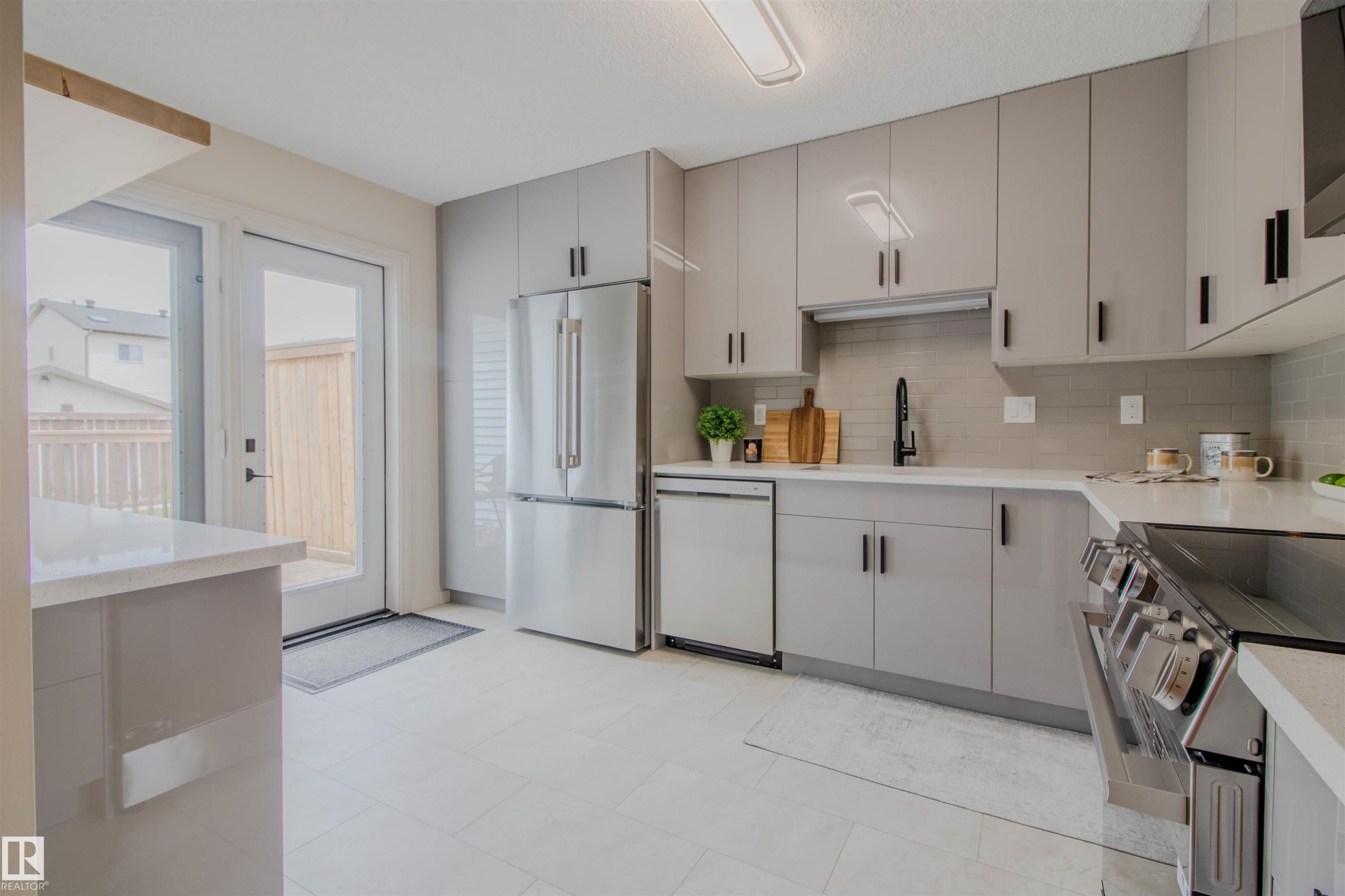 Modern kitchen featuring sleek gray cabinetry, stainless steel appliances, light-toned countertops, a full-height tile backsplash, and a glass-paned door leading to the exterior - 5935 189 Street, Edmonton, AB - Indoor Photo Showing Kitchen