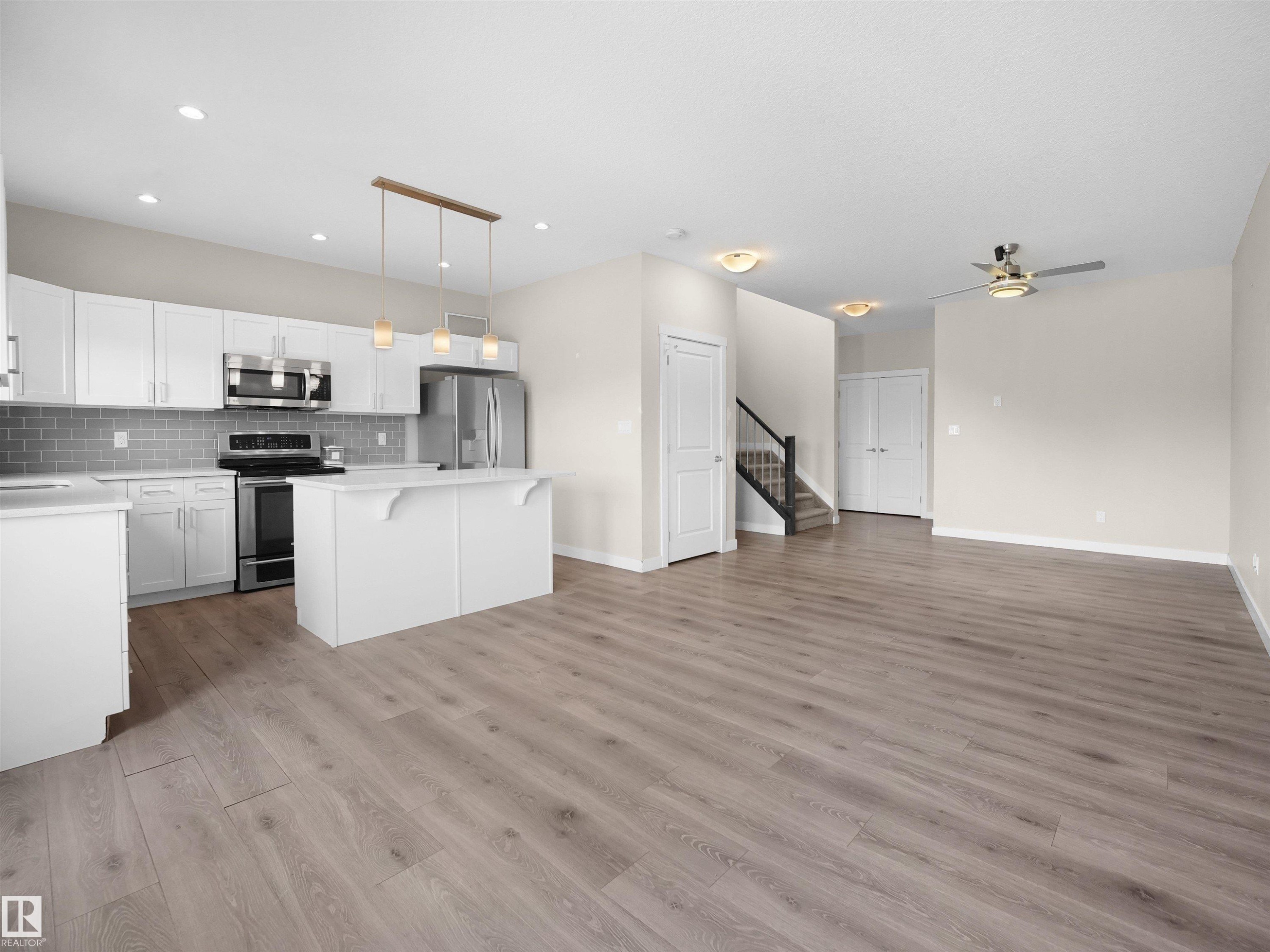 Open-concept kitchen and living area featuring wood-finish flooring, white cabinetry, stainless steel appliances, a kitchen island, and a staircase with dark banisters - 8921 217 Street Nw, Edmonton, AB - Indoor Photo Showing Kitchen