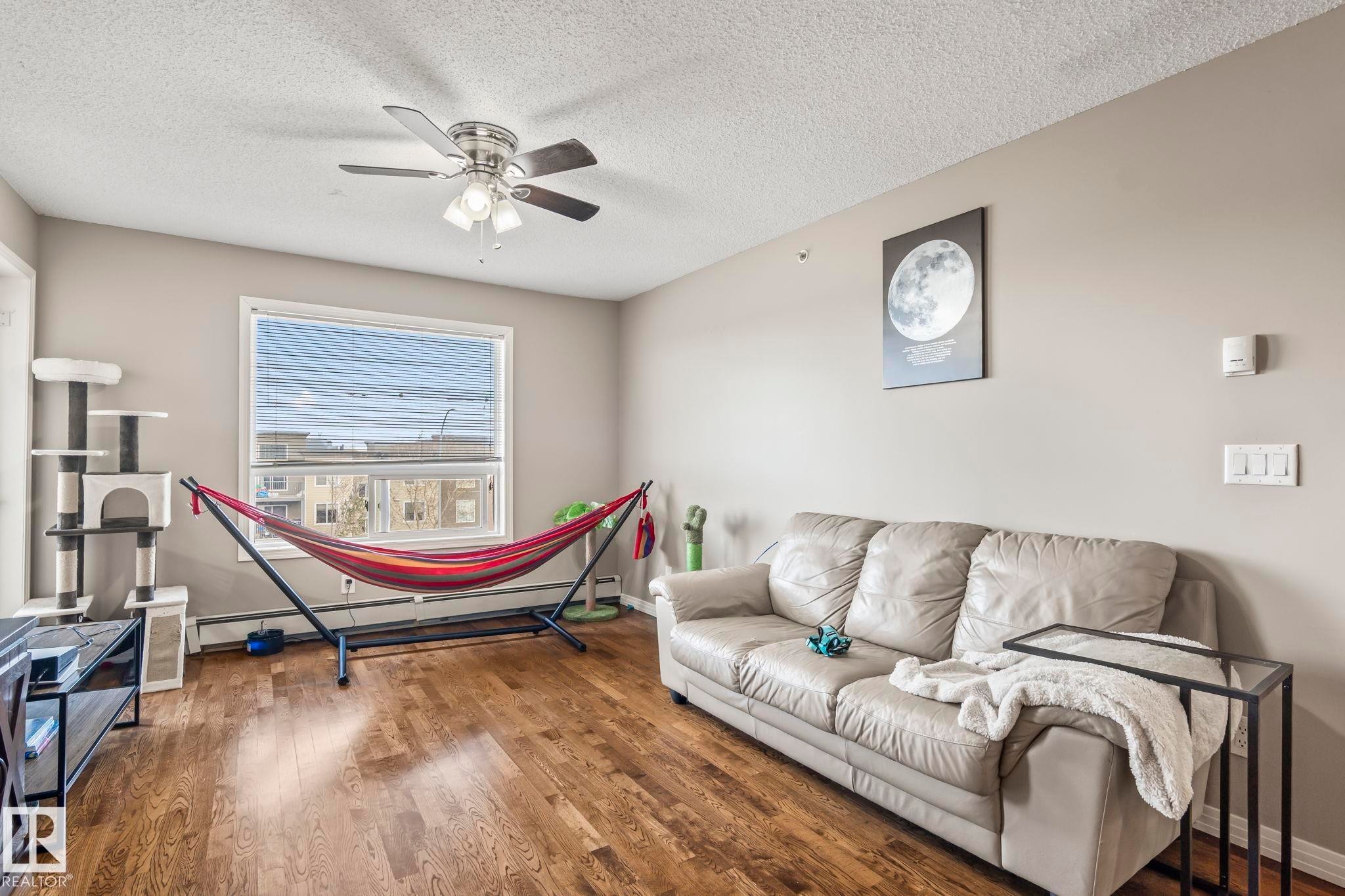 Spacious living area featuring wood-finish flooring, a large window with blinds, and a ceiling fan with integrated lighting - 412 5005 165 Avenue Nw, Edmonton, AB - Indoor Photo Showing Living Room