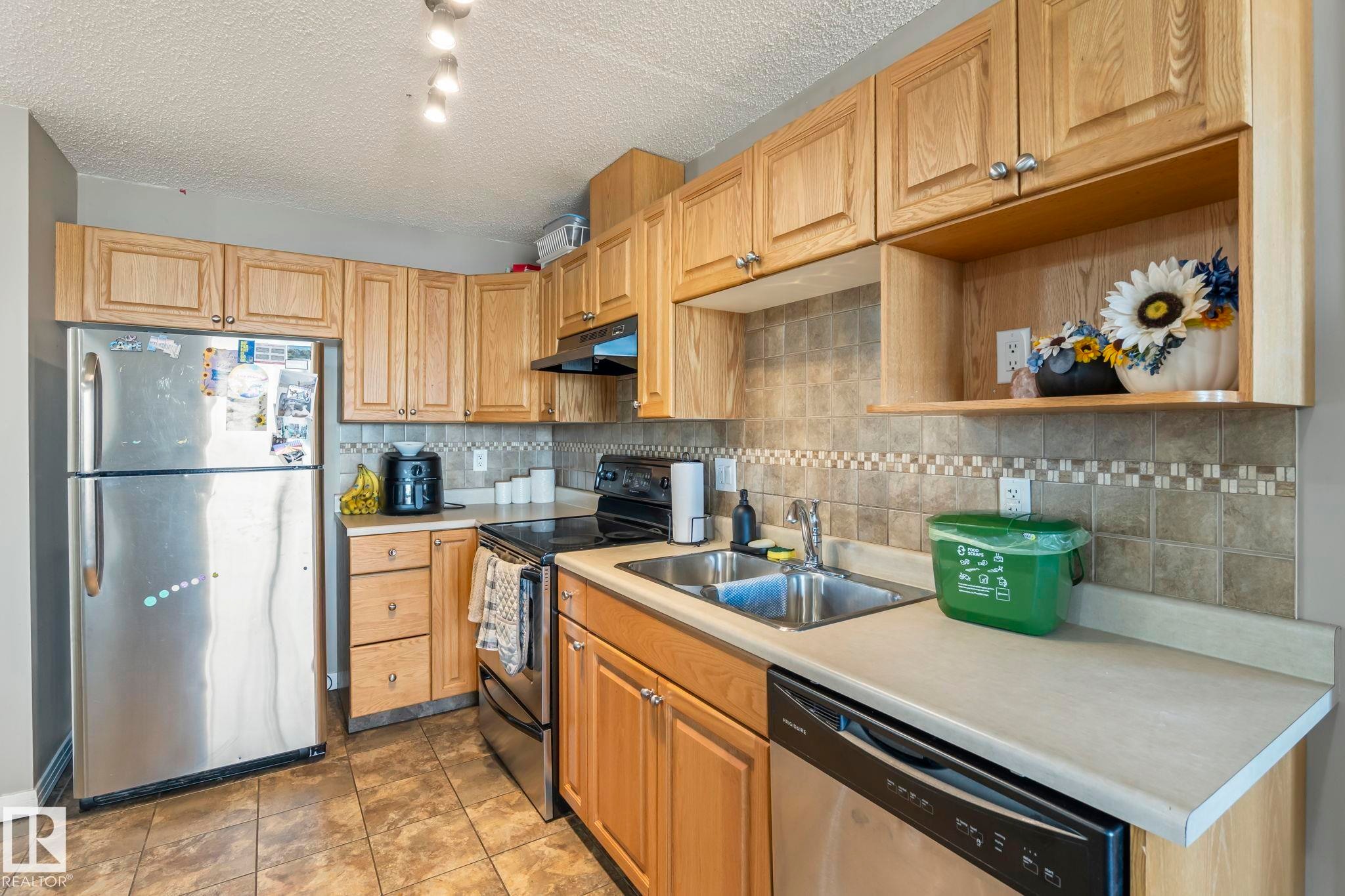 Kitchen featuring wood cabinetry, stainless steel appliances, a double basin sink, light-toned countertops, and a tiled backsplash with a decorative accent band - 412 5005 165 Avenue Nw, Edmonton, AB - Indoor Photo Showing Kitchen With Double Sink