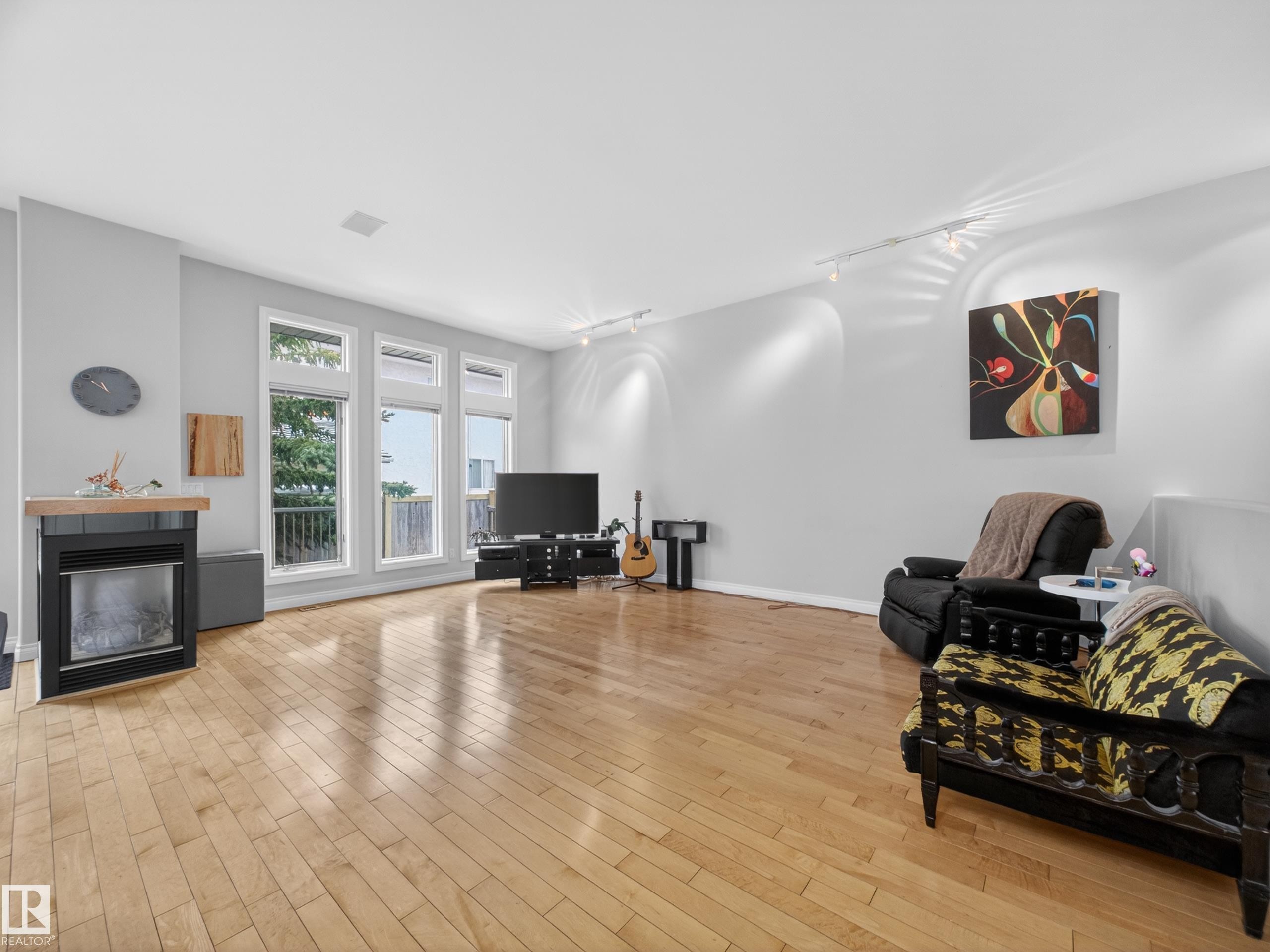 Spacious living area featuring light wood-finish flooring, a wall-mounted fireplace with wood mantel, a series of large windows, and recessed ceiling lighting - 569 Butterworth Way, Edmonton, AB - Indoor Photo Showing Living Room With Fireplace