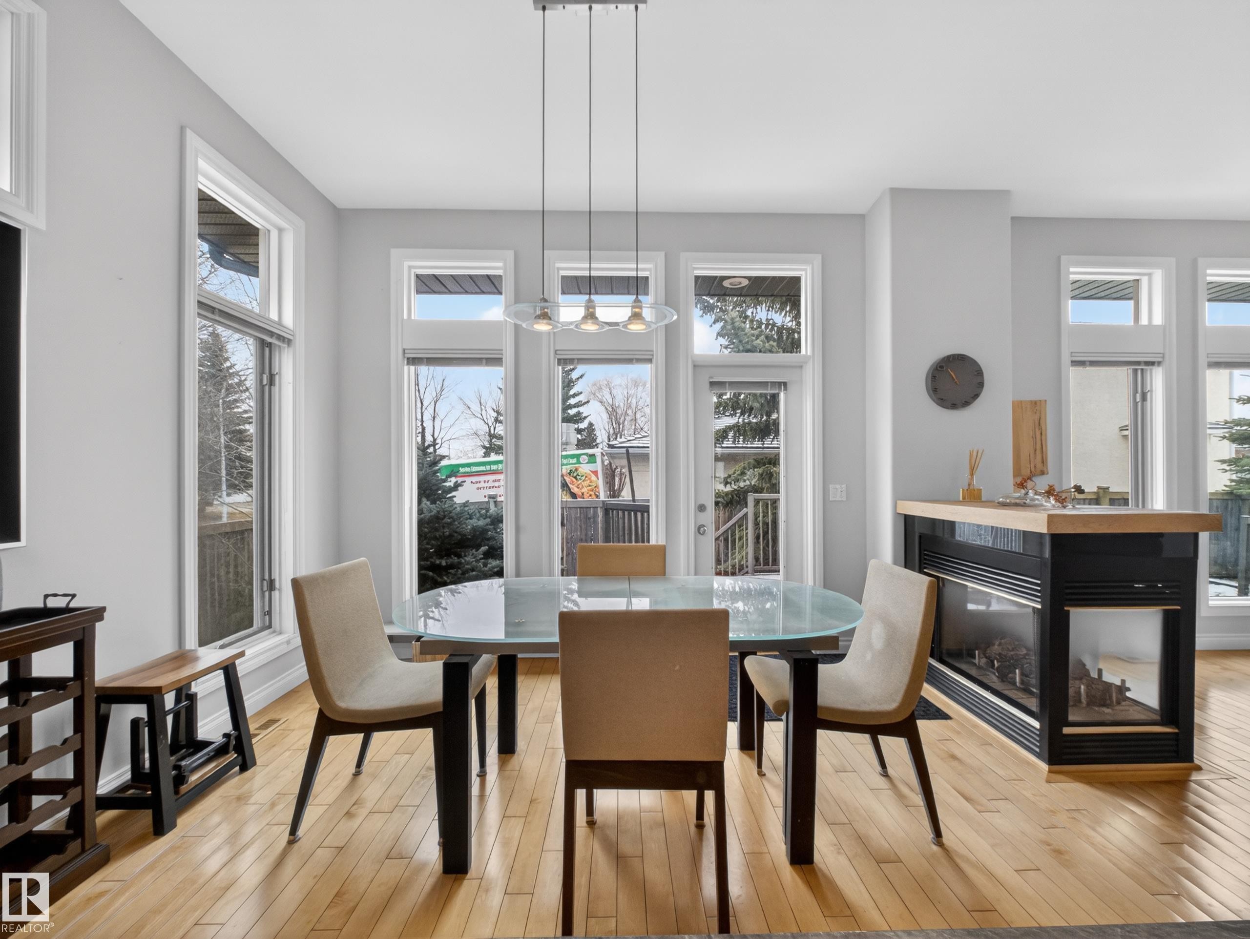 Light-filled dining area featuring wood-finish flooring, a contemporary linear chandelier, multiple large windows, and a built-in double-sided fireplace with a wood-finish top - 569 Butterworth Way, Edmonton, AB - Indoor Photo Showing Dining Room