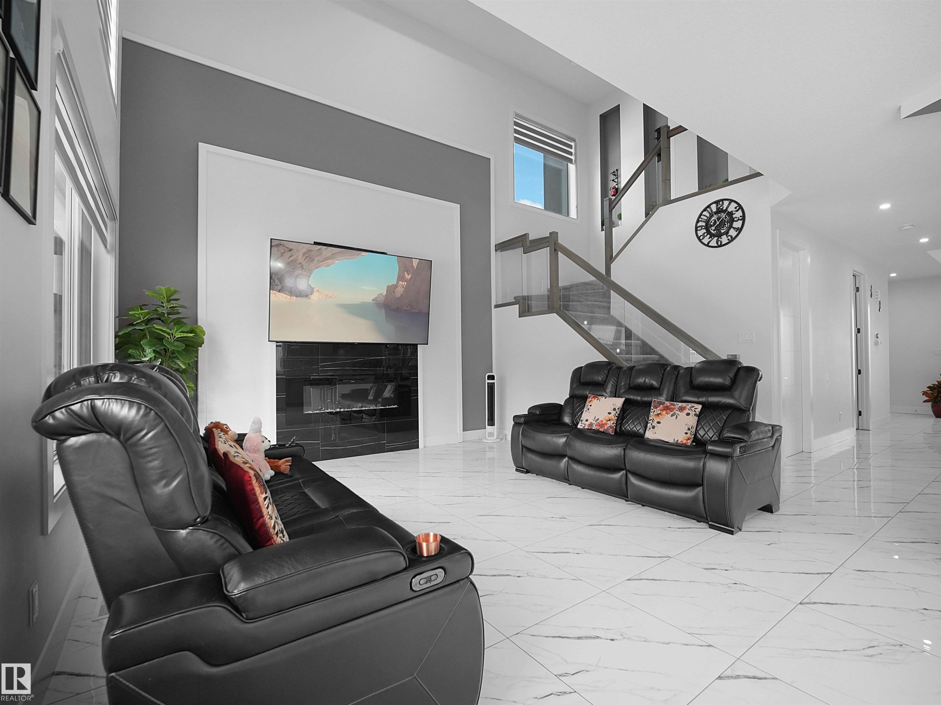 Open-concept living space with large format white tile flooring, a contemporary fireplace feature wall with a mounted television, a modern staircase with glass railing, and recessed lighting throughout - 1208 25 St Nw, Edmonton, AB - Indoor