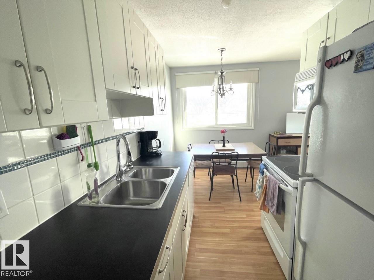 Bright kitchen featuring extensive white cabinetry, a double basin stainless steel sink, and dark countertops - #303 12223 82 St Nw, Edmonton, AB - Indoor Photo Showing Kitchen With Double Sink