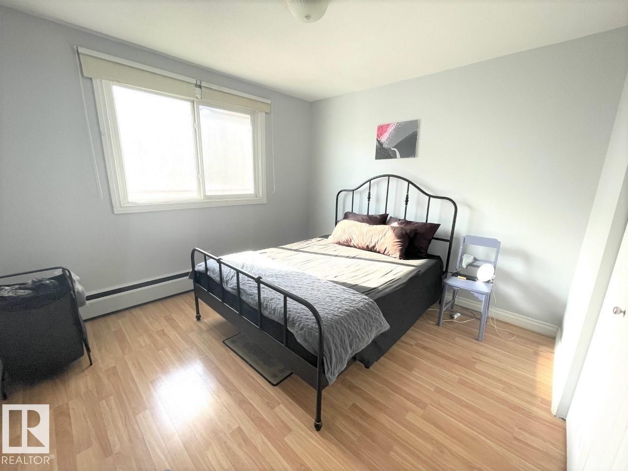 Spacious bedroom featuring light wood-finish flooring, a large window, and a baseboard heating unit - #303 12223 82 St Nw, Edmonton, AB - Indoor Photo Showing Bedroom