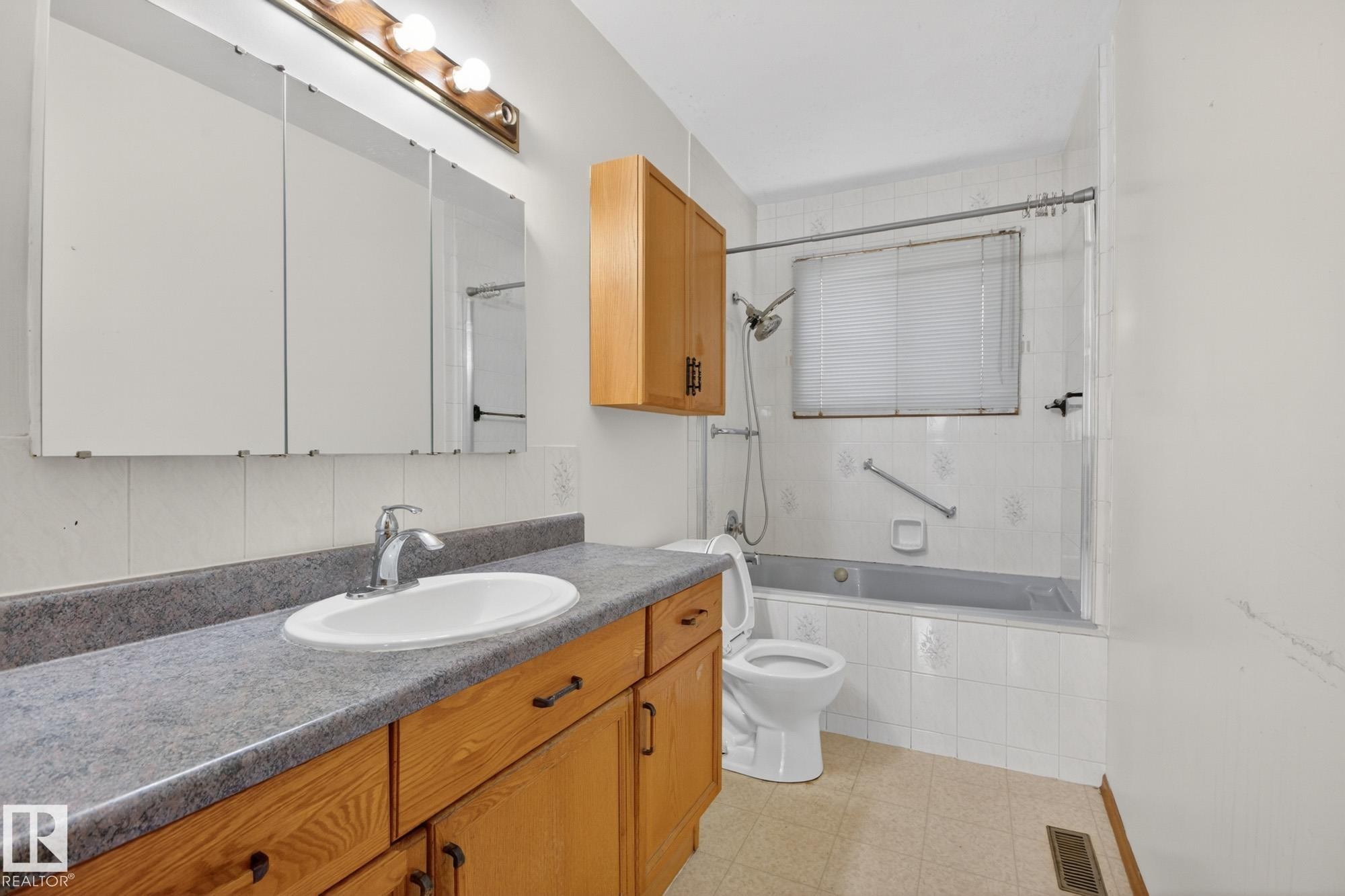 Bathroom featuring a wood vanity with a gray countertop and integrated sink, a three-panel mirrored medicine cabinet, a bathtub with a showerhead and white tile surround, and an overhead light fixture - 11901 42 Street Nw, Edmonton, AB - Indoor Photo Showing Bathroom